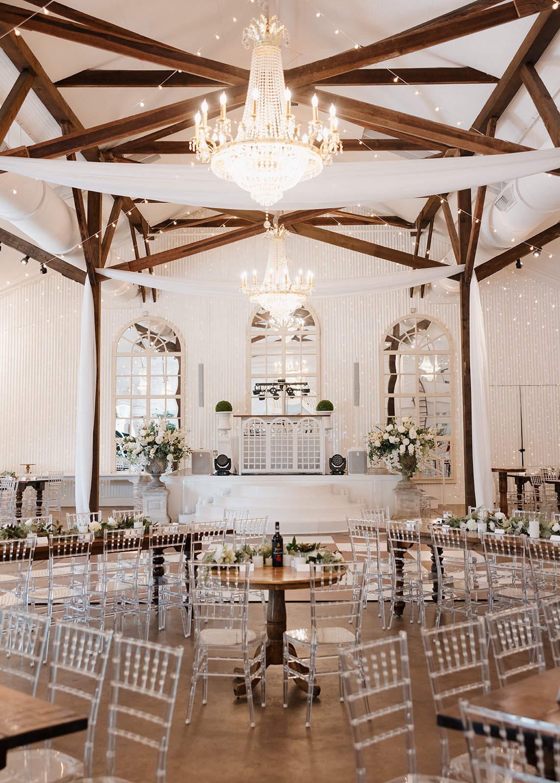 Elegant wedding reception hall decorated with white flowers, chandeliers, and string lights, featuring clear chairs, wooden tables, and a stage with DJ equipment.