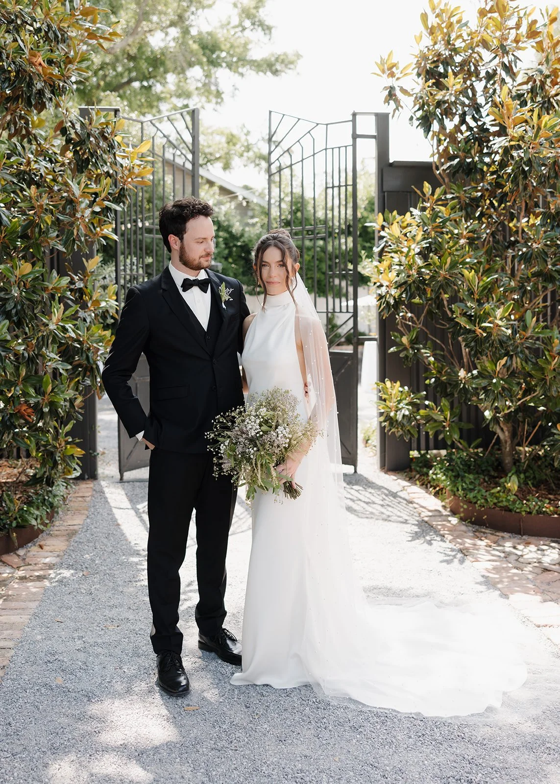 A Pensacola bride and groom standing outdoors, with the bride in a white gown holding a bouquet, and the groom in a black suit with a bow tie, in front of a gate surrounded by greenery.