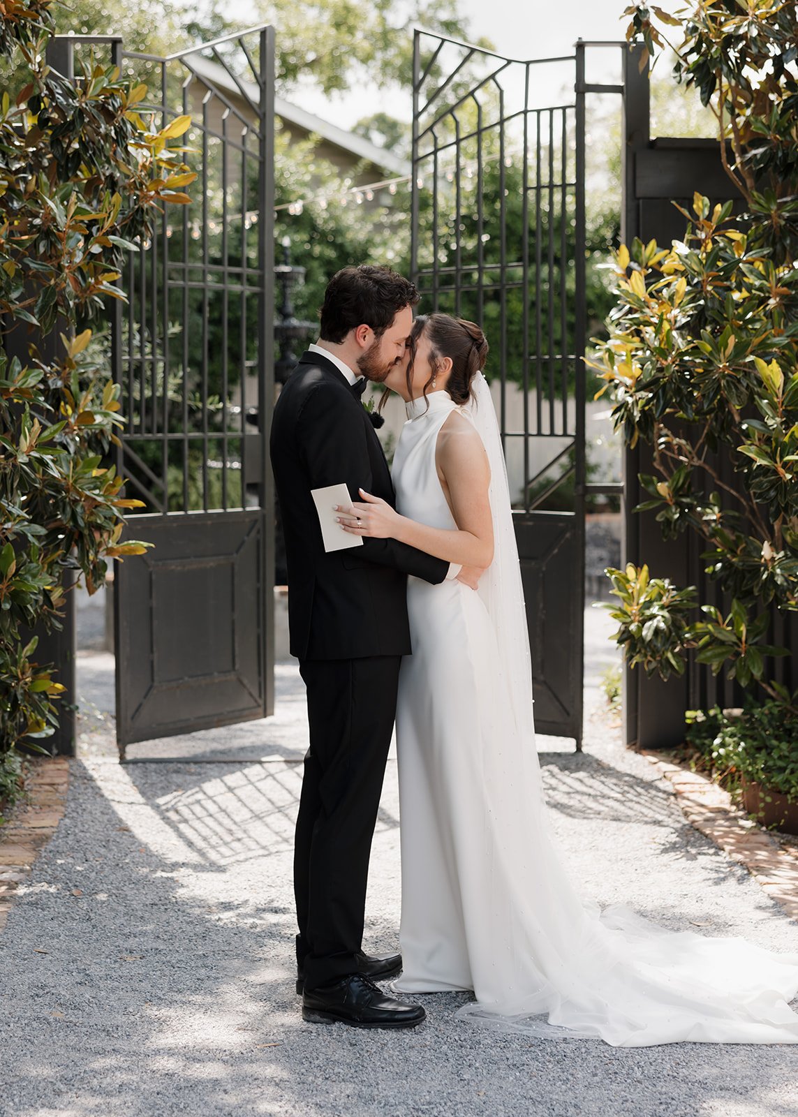 A newlywed couple sharing a kiss at their Pensacola wedding ceremony outdoors, with a black metal gate and greenery in the background.