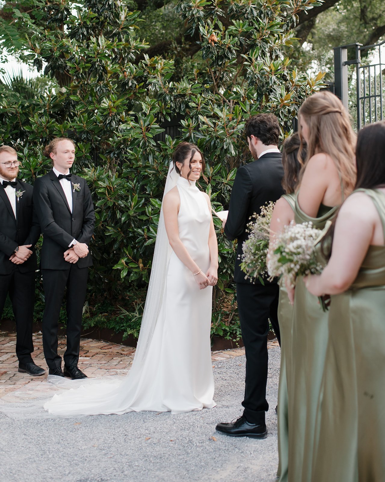 A wedding ceremony outdoors with a bride in a white gown and veil, groom in a black suit, standing before an officiant. Bridesmaids in matching olive green dresses hold flowers, groomsmen in black suits stand nearby, and a green leafy backdrop surrou