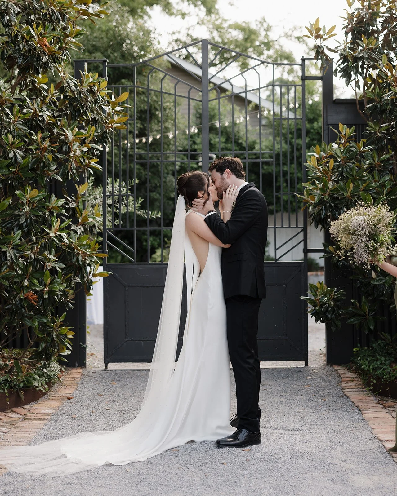 A bride and groom sharing a kiss at their wedding ceremony outdoors, with greenery and large black gate in the background.