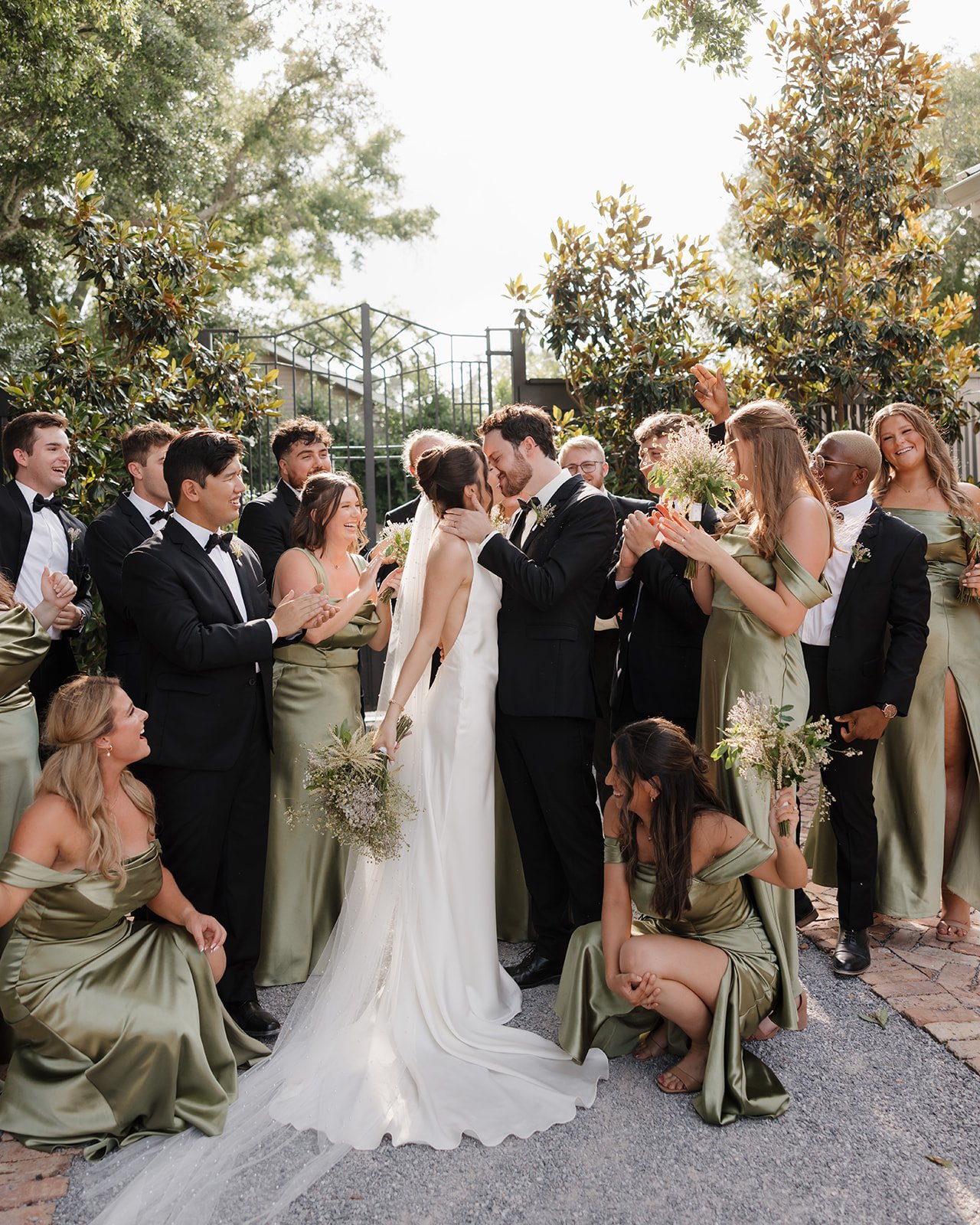 A Pensacola wedding celebration with the bride and groom kissing in the center, surrounded by friends and family who are clapping and cheering outside under trees.