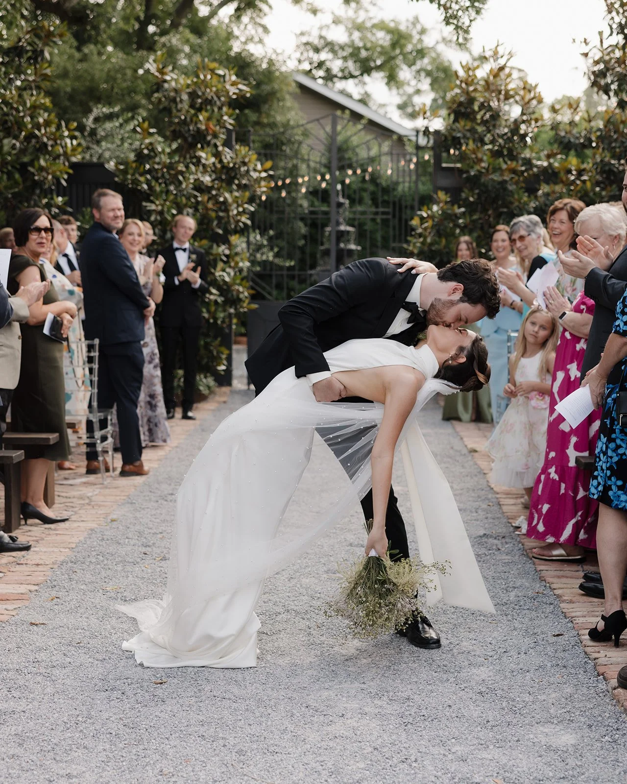 A Pensacola bride and groom share a kiss during their wedding ceremony, with the groom dipping the bride. The bride holds a bouquet, and guests are clapping and smiling around them outdoors.

