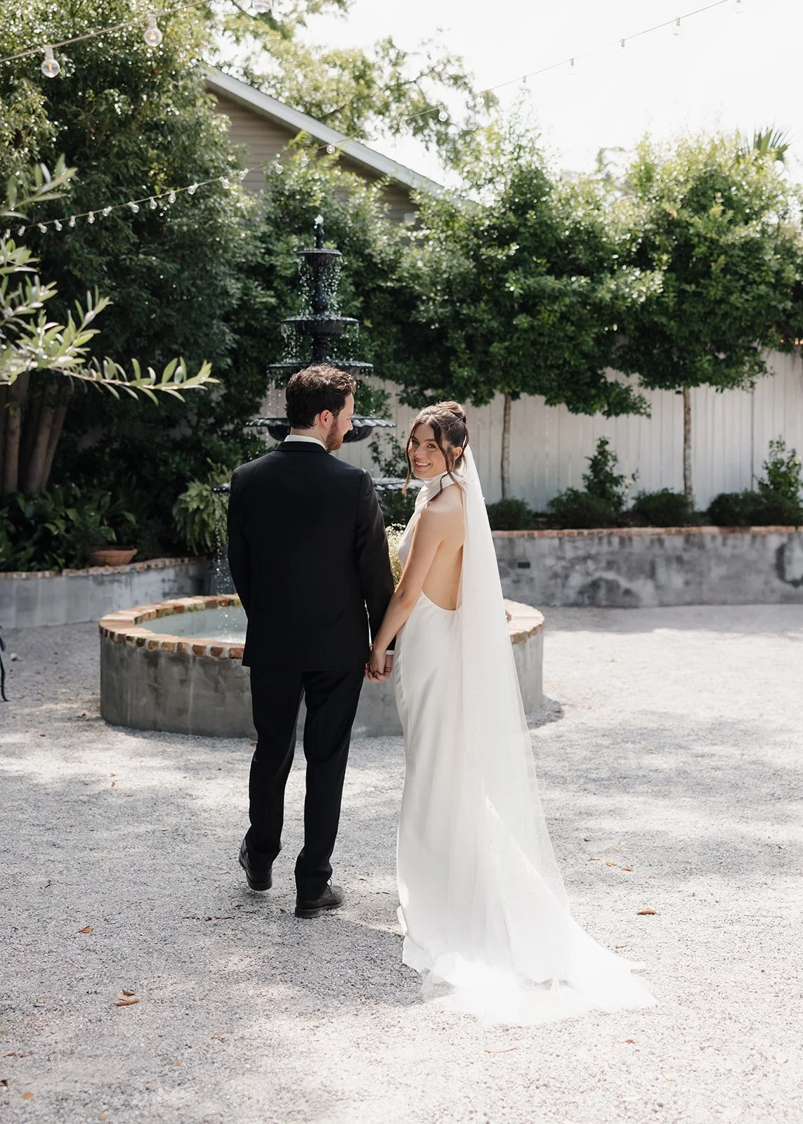 A bride and groom holding hands, standing outdoors near a fountain with trees and string lights overhead, during daytime at a Pensacola Wedding Venue.