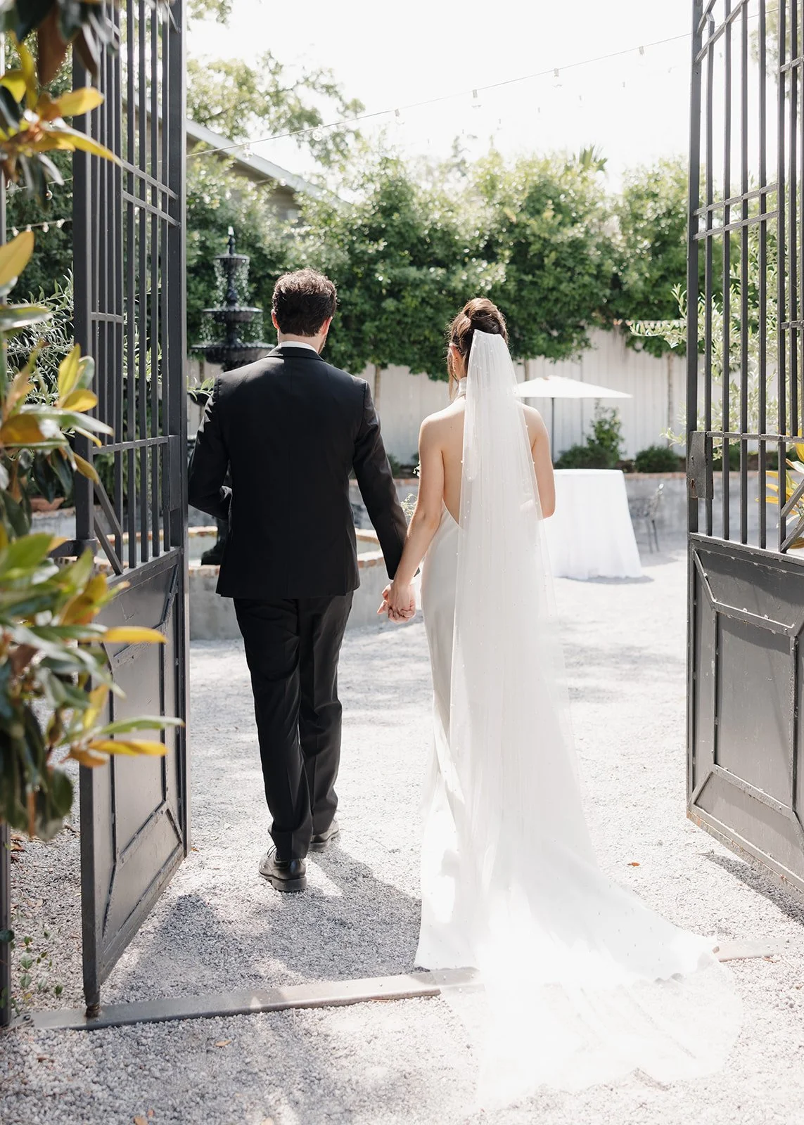 A bride and groom walking hand in hand through a gate, outdoors, with trees and string lights overhead, a fountain and table with an umbrella visible in the background.
