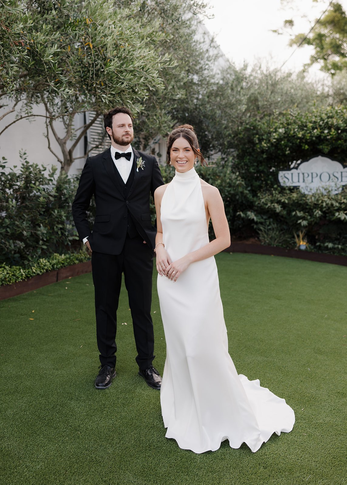 A bride and groom in wedding attire standing outdoors on a green lawn, with trees and bushes in the background. The bride is smiling, wearing a white sleeveless wedding dress with a high neckline, and the groom is standing slightly behind her, wearin