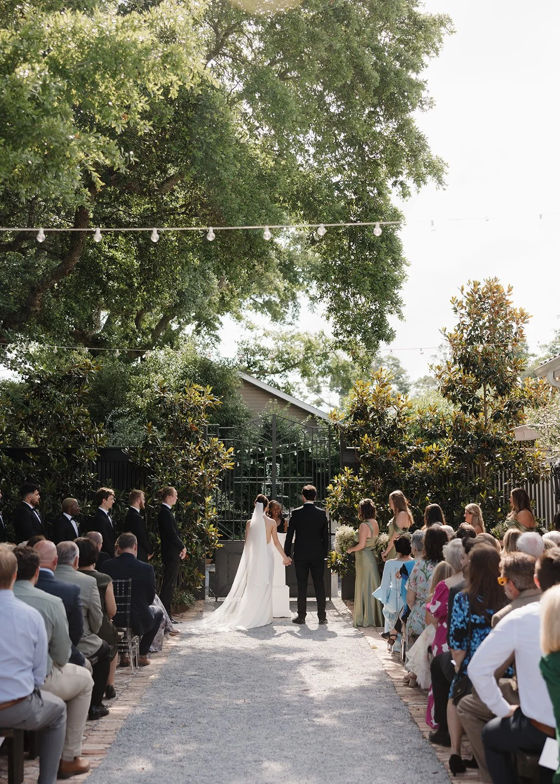 Pensacola Wedding ceremony outdoors with bride and groom holding hands, surrounded by guests seated on either side, under string lights and trees.