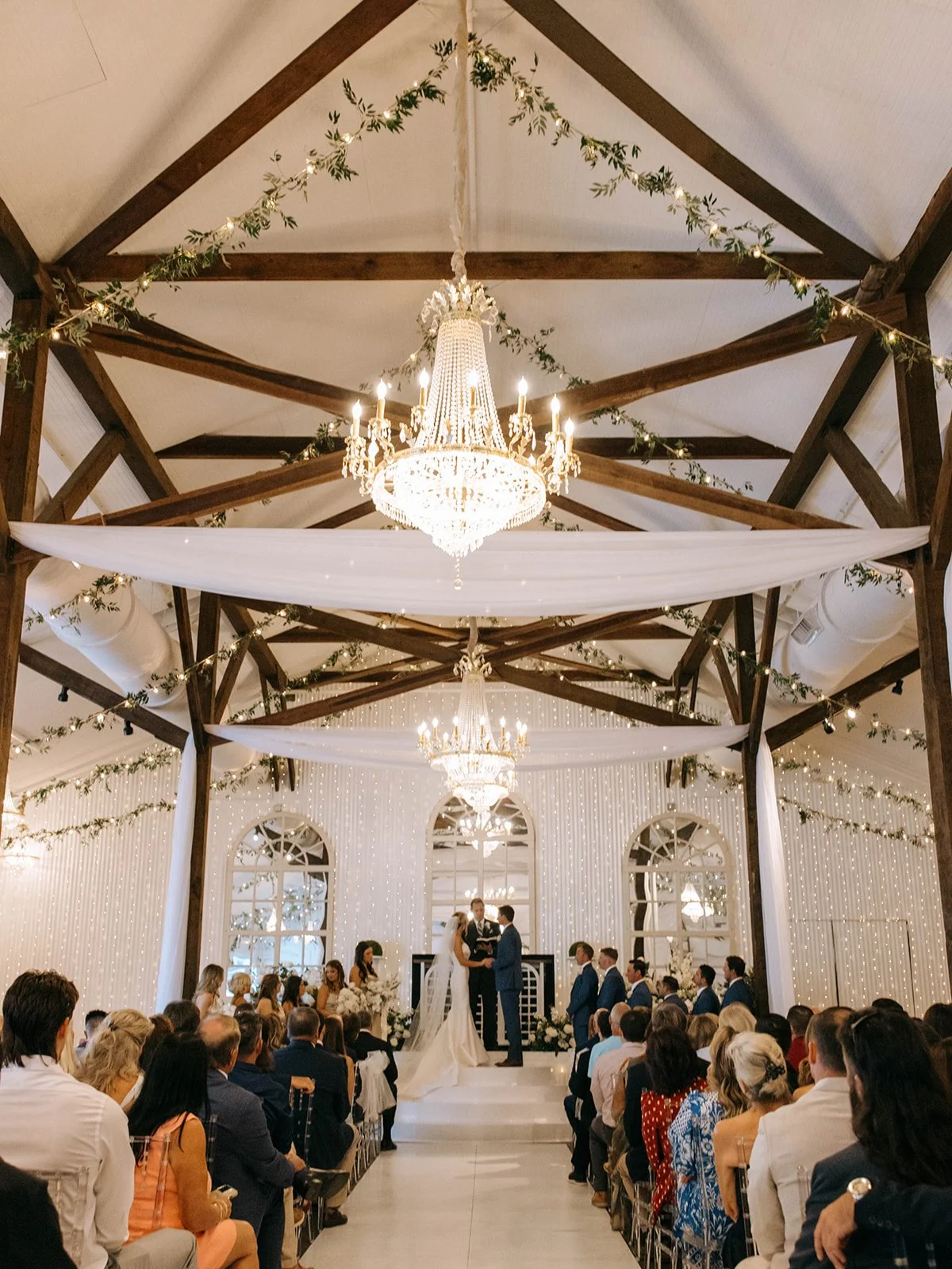 A wedding ceremony with a bride and groom holding hands under ornate chandeliers, with guests seated on either side in a decorated venue.