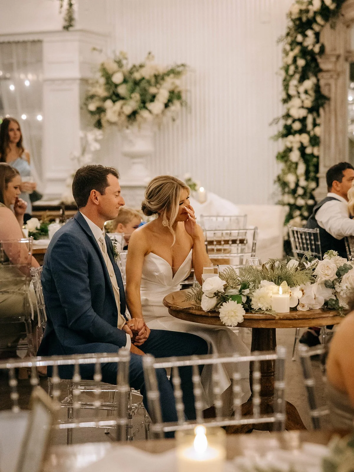 A bride and groom sitting at a wedding reception table with white flowers and candles, surrounded by guests, in an elegant decorated indoor venue.
