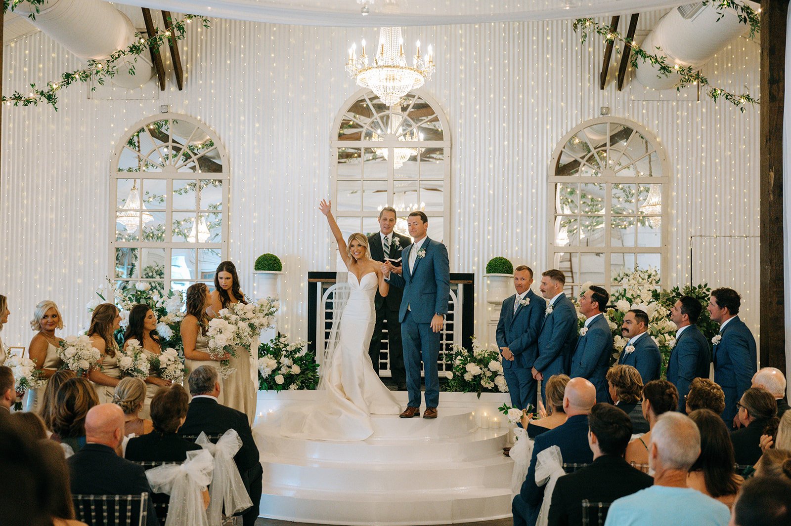 Bride and groom exchanging vows during wedding ceremony in a decorated indoor venue with chandeliers, fairy lights, large windows, and floral arrangements.