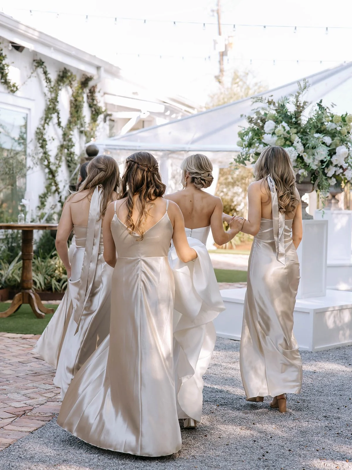 Group of bridesmaids in champagne-colored satin dresses walking outdoors during a wedding celebration, with large floral arrangements and white structures in the background.