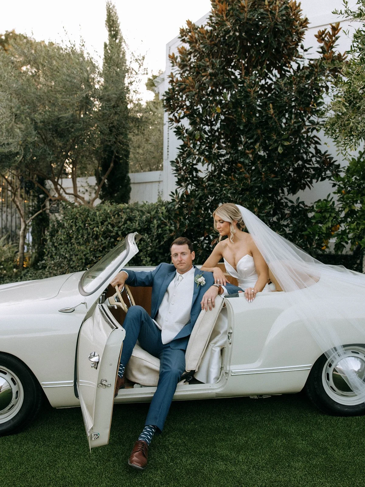 Bride and groom sitting in a vintage white convertible car with the bride leaning over the groom's shoulder, surrounded by greenery and trees.