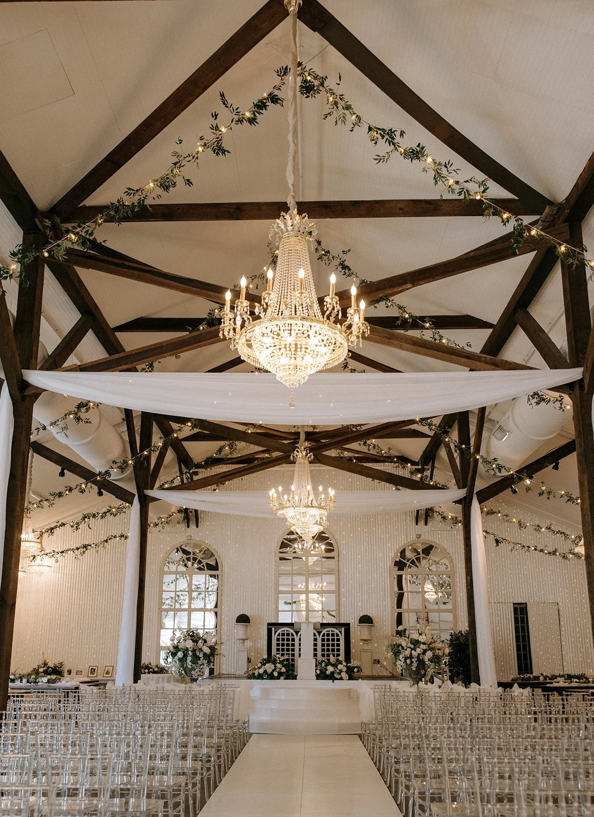 Elegant Pensacola wedding ceremony venue with chandeliers, white drapes, floral arrangements, and rows of clear chairs.