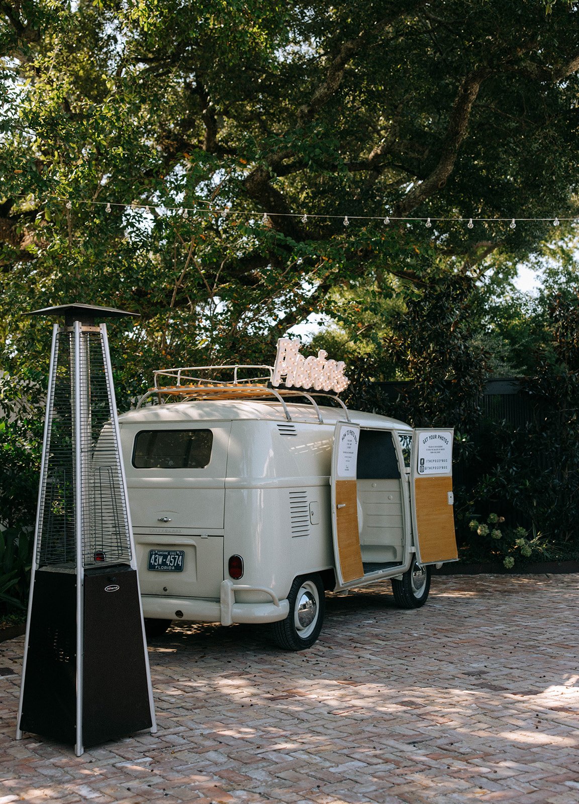 White vintage camper van with open door, parked on brick pavement under large green tree, with a sign on top that says 'Andie', and a tall outdoor heater nearby.