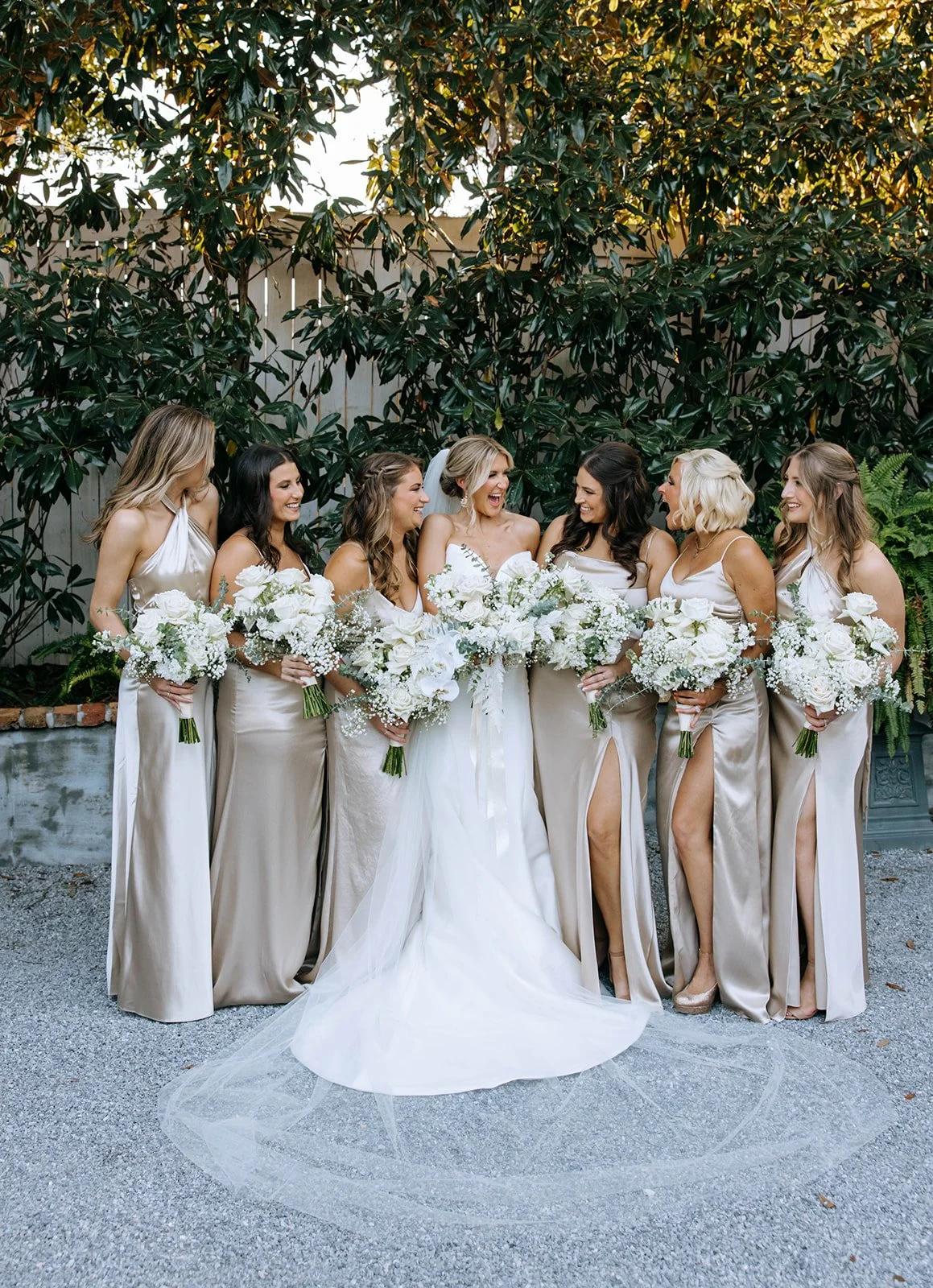Group of women, including a bride and bridesmaids, standing together outdoors, holding white bouquets, and laughing with greenery background.