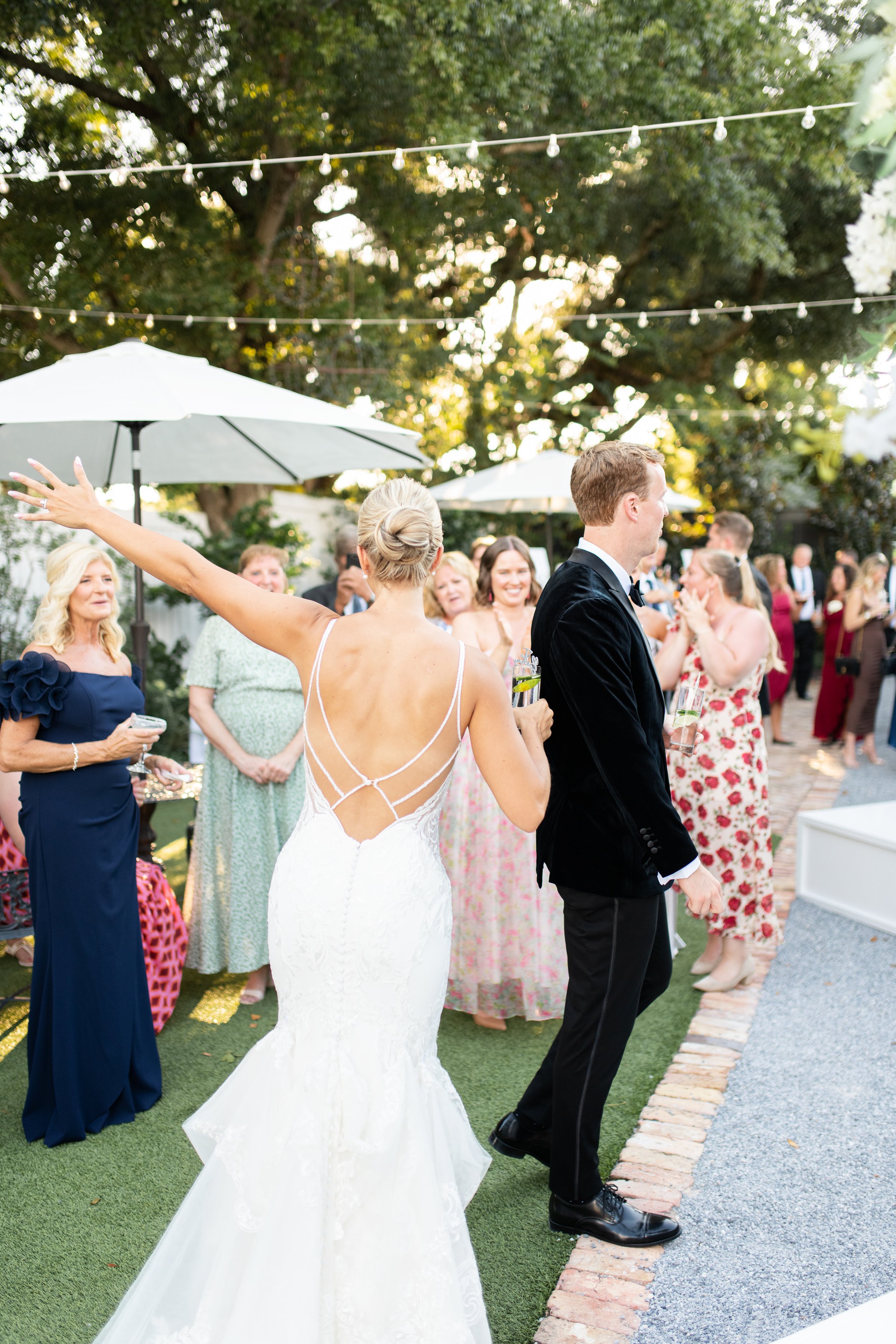 Bride with her back to the camera, wearing a white wedding gown with an intricate back design, gesturing with her arms at a wedding celebration outdoors. Groomsman in a black tuxedo standing nearby. Guests in colorful dresses holding drinks, standing