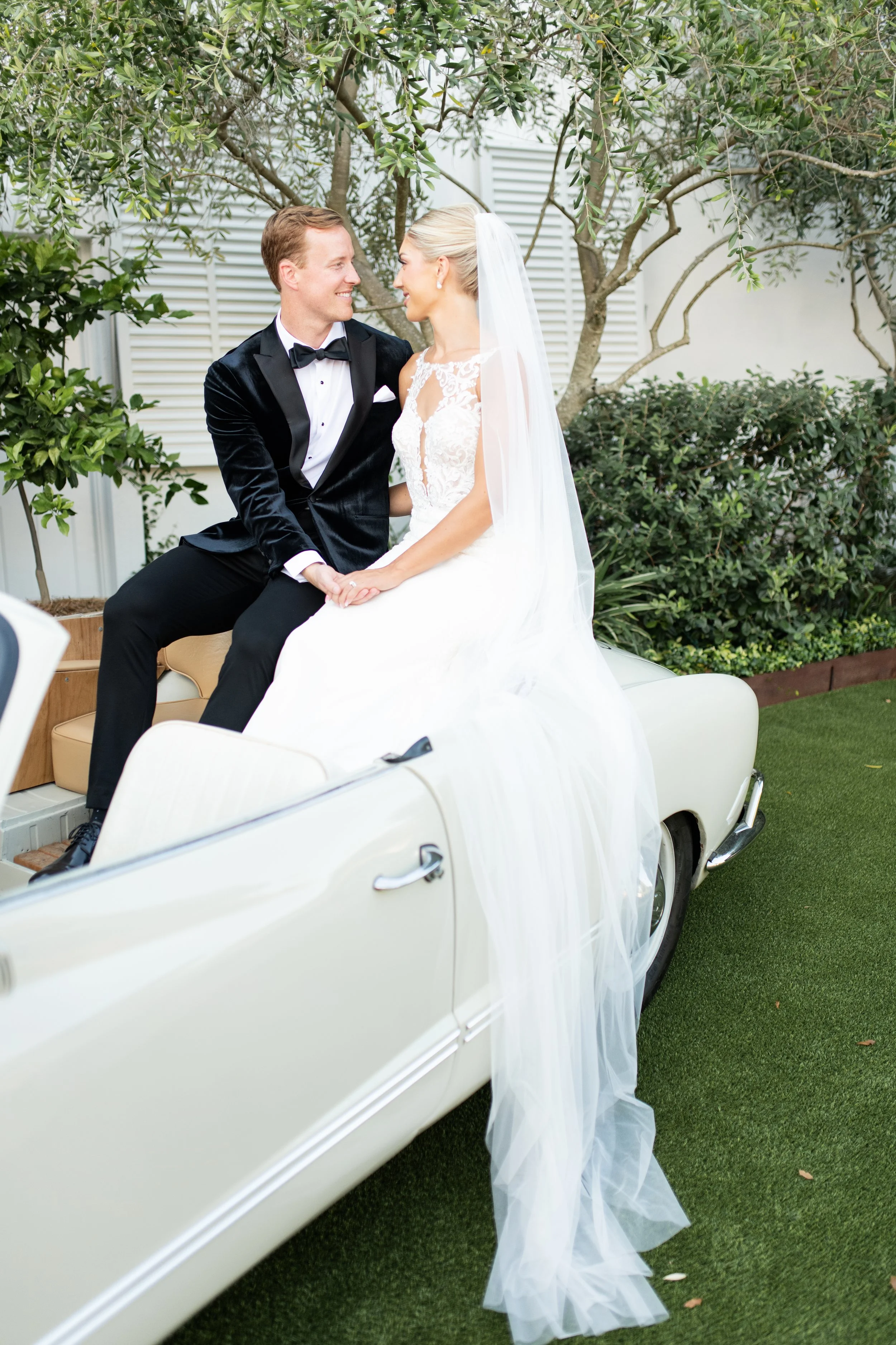 A bride and groom sitting together on a vintage white car outdoors, surrounded by greenery, smiling at each other during their wedding.