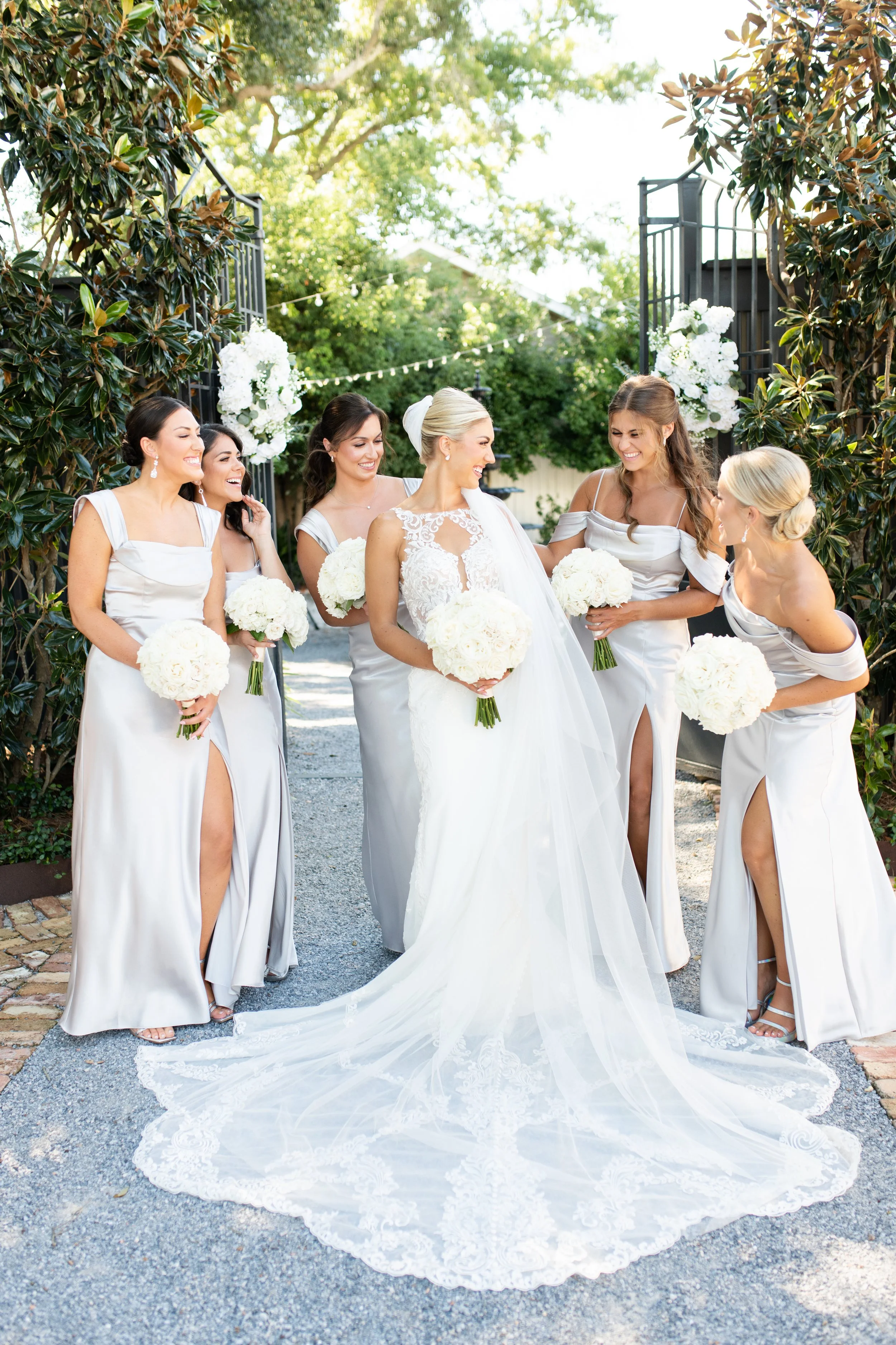 Bride in white wedding dress with lace details and veil, surrounded by five bridesmaids in matching off-shoulder satin dresses with high slits, holding white bouquets, standing under decorative archway with greenery and flowers on a sunny day.