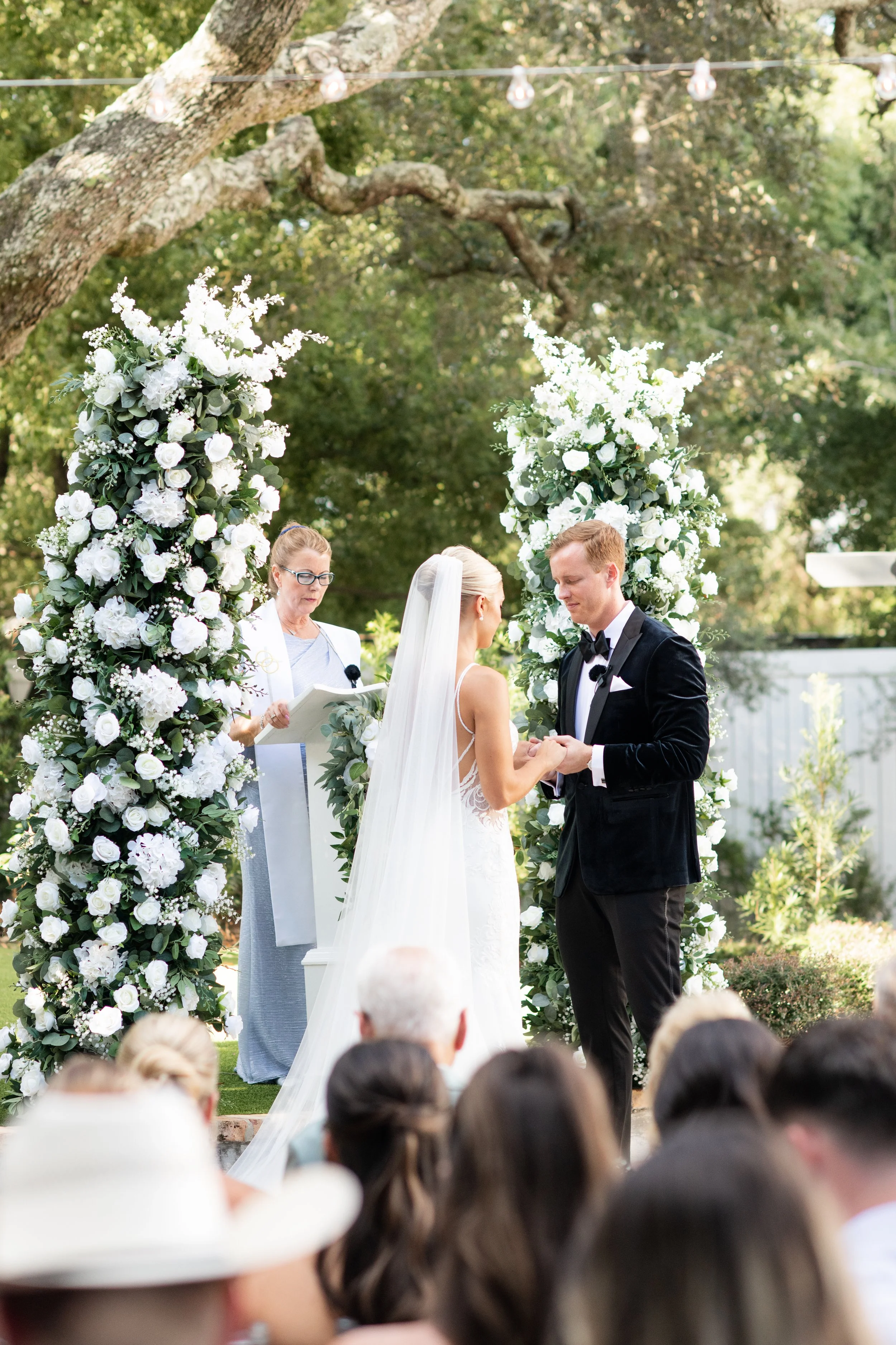 A Pensacola wedding ceremony outdoors with a bride and groom exchanging rings under a floral arch, with guests watching.