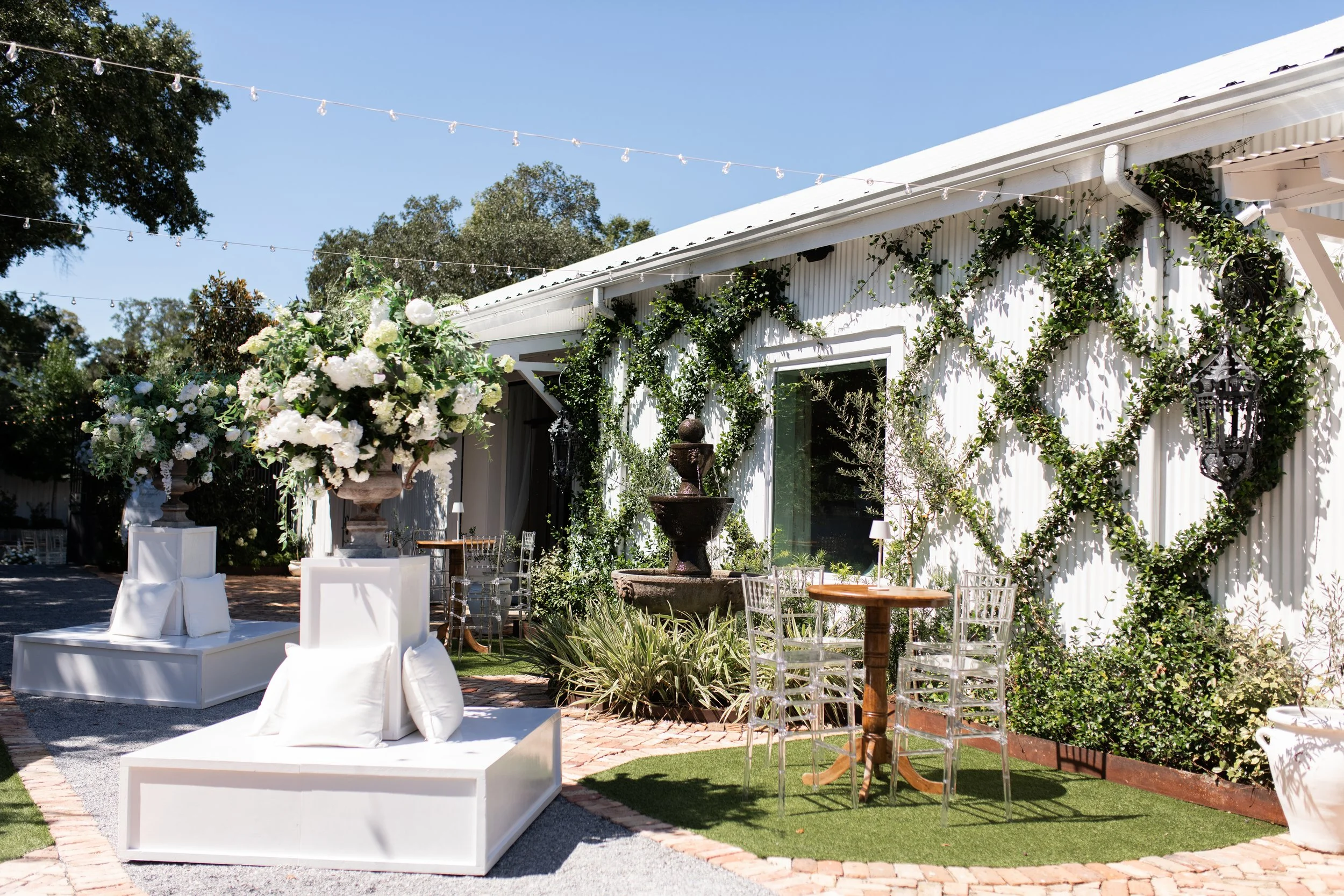 Outdoor event space with white furniture, large floral arrangements, a fountain, and string lights, surrounded by greenery and a white building with green vines.
