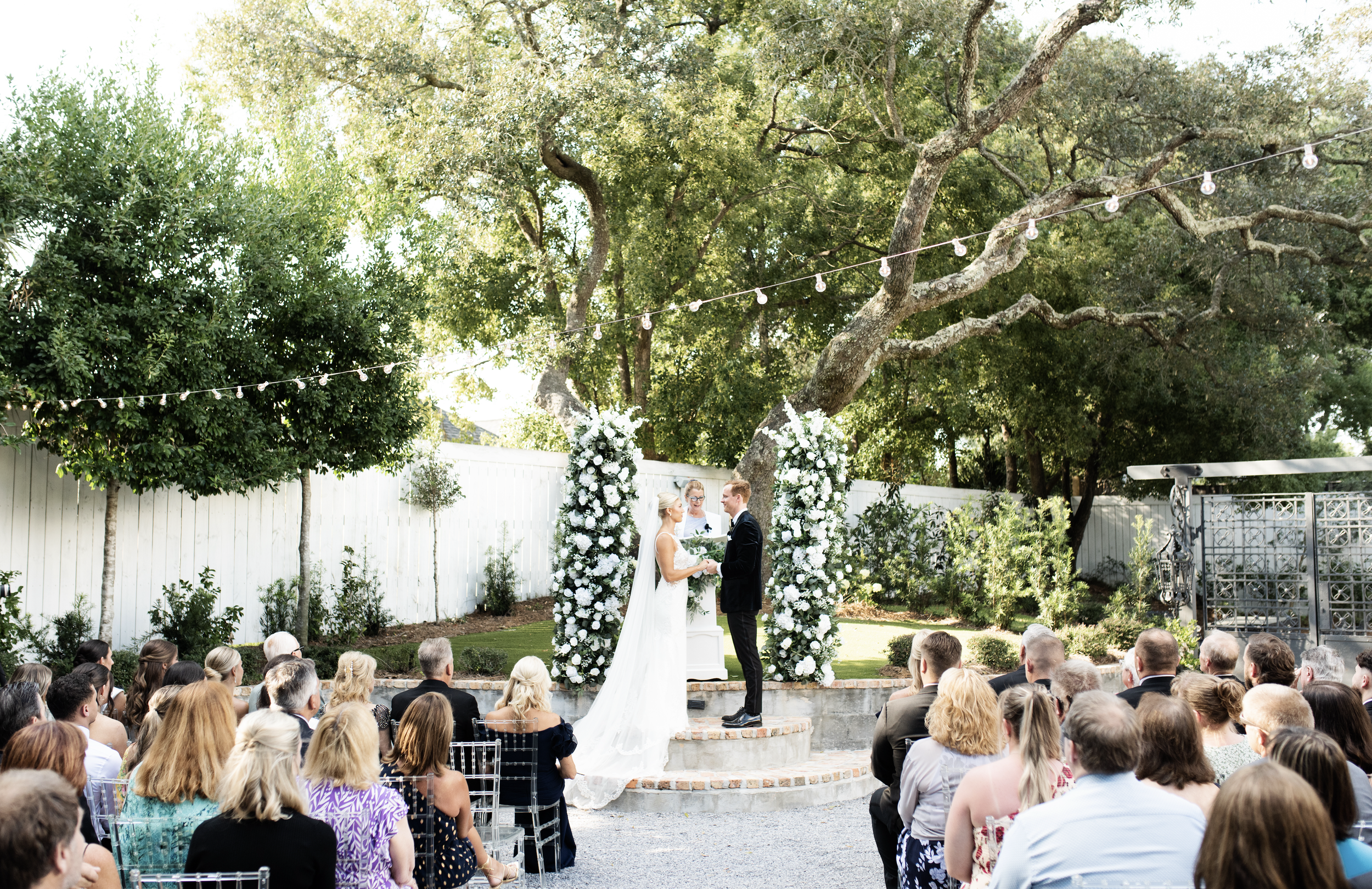 A wedding ceremony taking place outdoors under a large tree, with a bride and groom standing under an arch of white flowers, facing each other and holding hands, while an officiant stands behind them surrounded by guests seated on chairs.