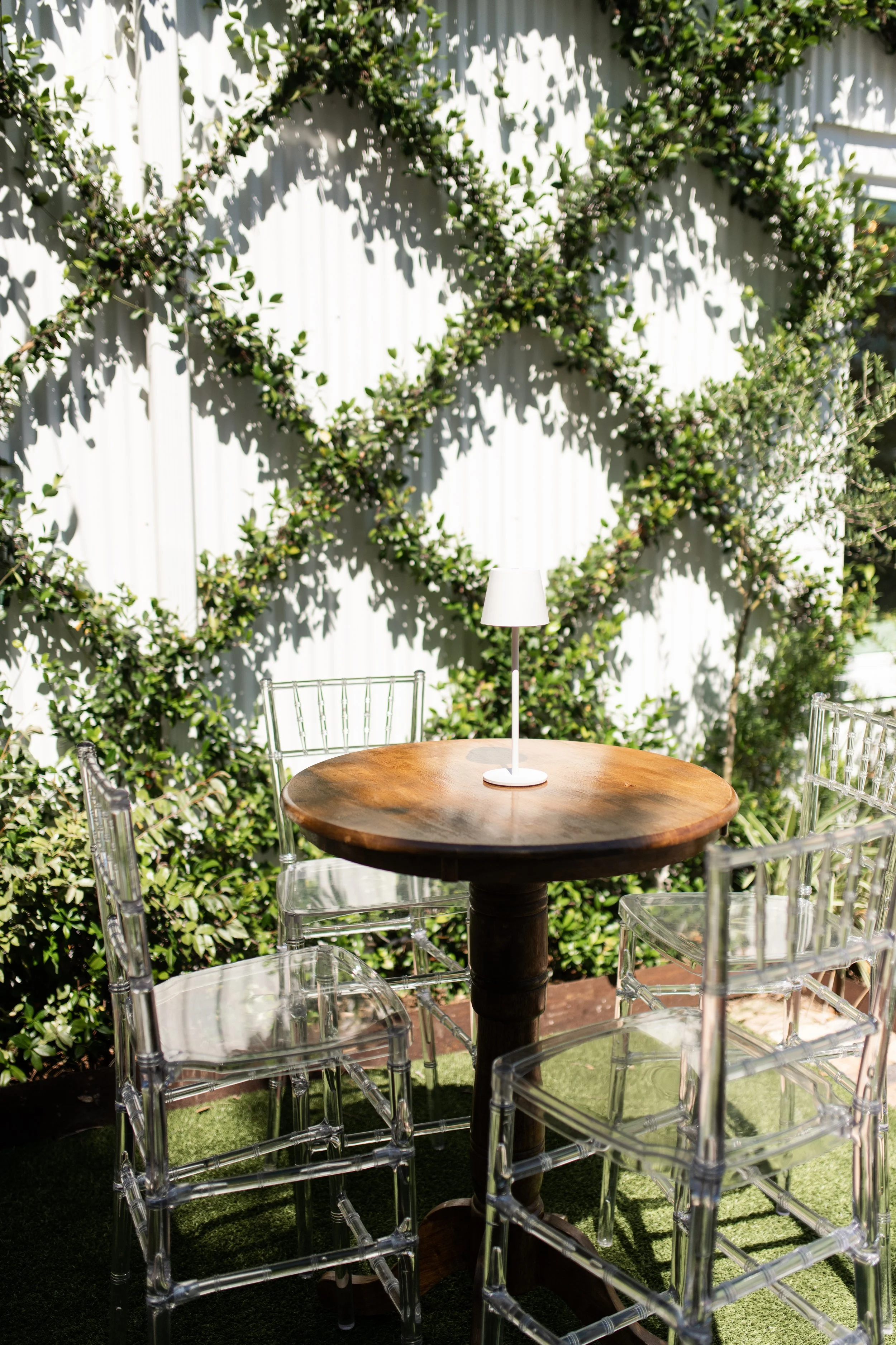 A round wooden table with a small white lamp on it, surrounded by four transparent ghost chairs, set outdoors against a white wall with green climbing plants creating a diamond pattern.