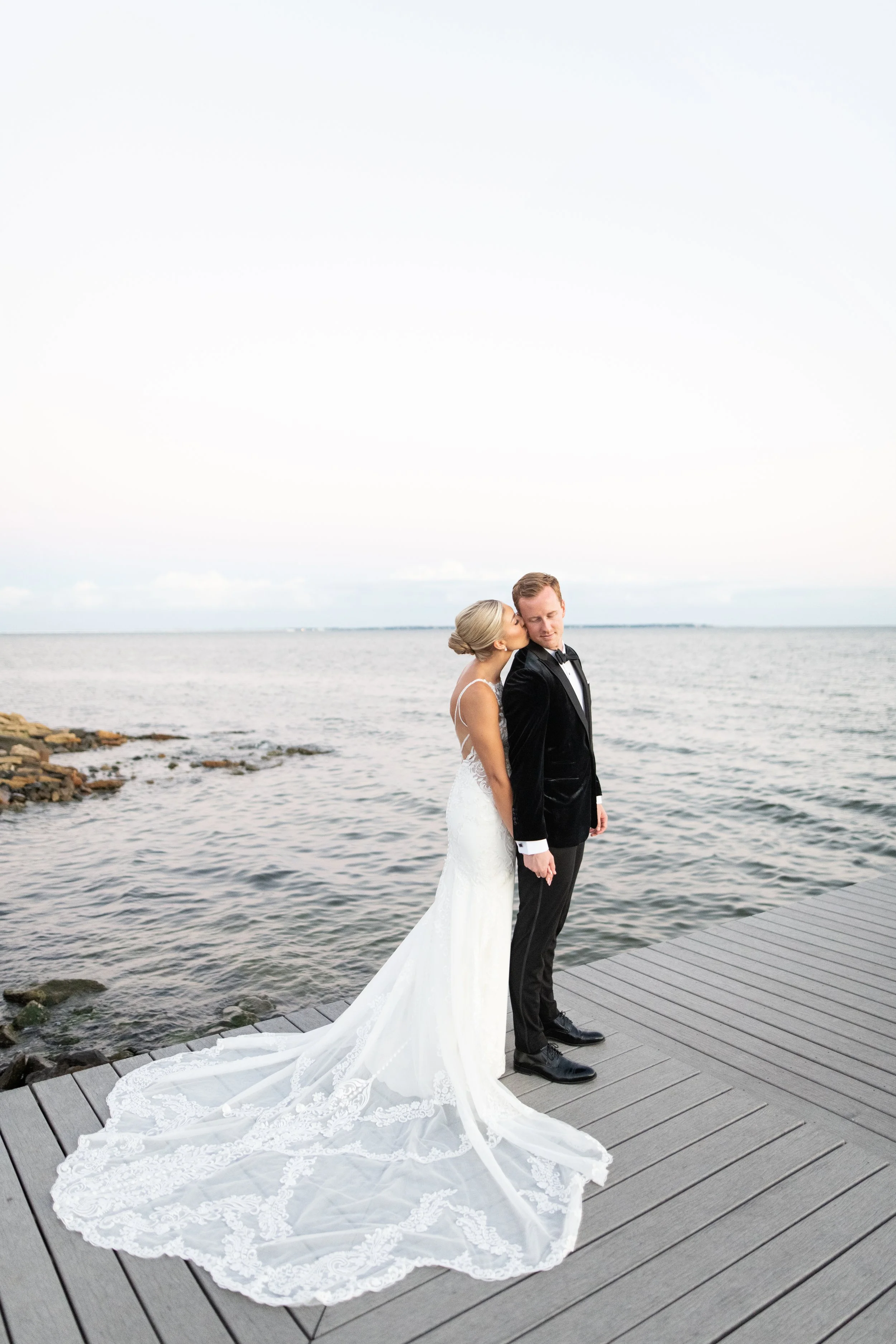 Bride and groom standing on a wooden dock beside the water, with the bride kissing the groom on the cheek, during their wedding.