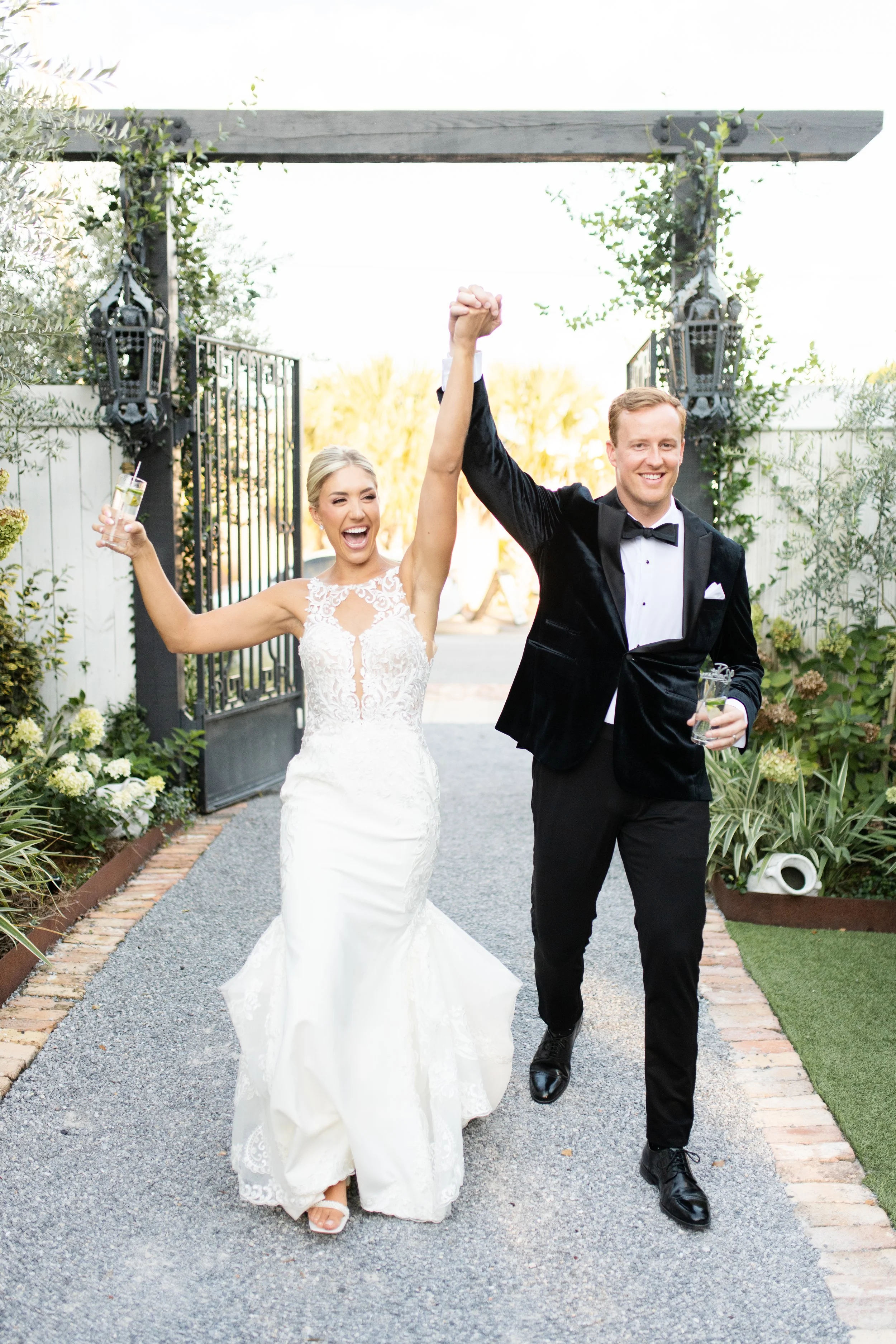 A newlywed couple celebrating, with the bride in a lace wedding dress and the groom in a tuxedo, walking under an outdoor archway. They are smiling, holding drinks, and raising their hands in victory.