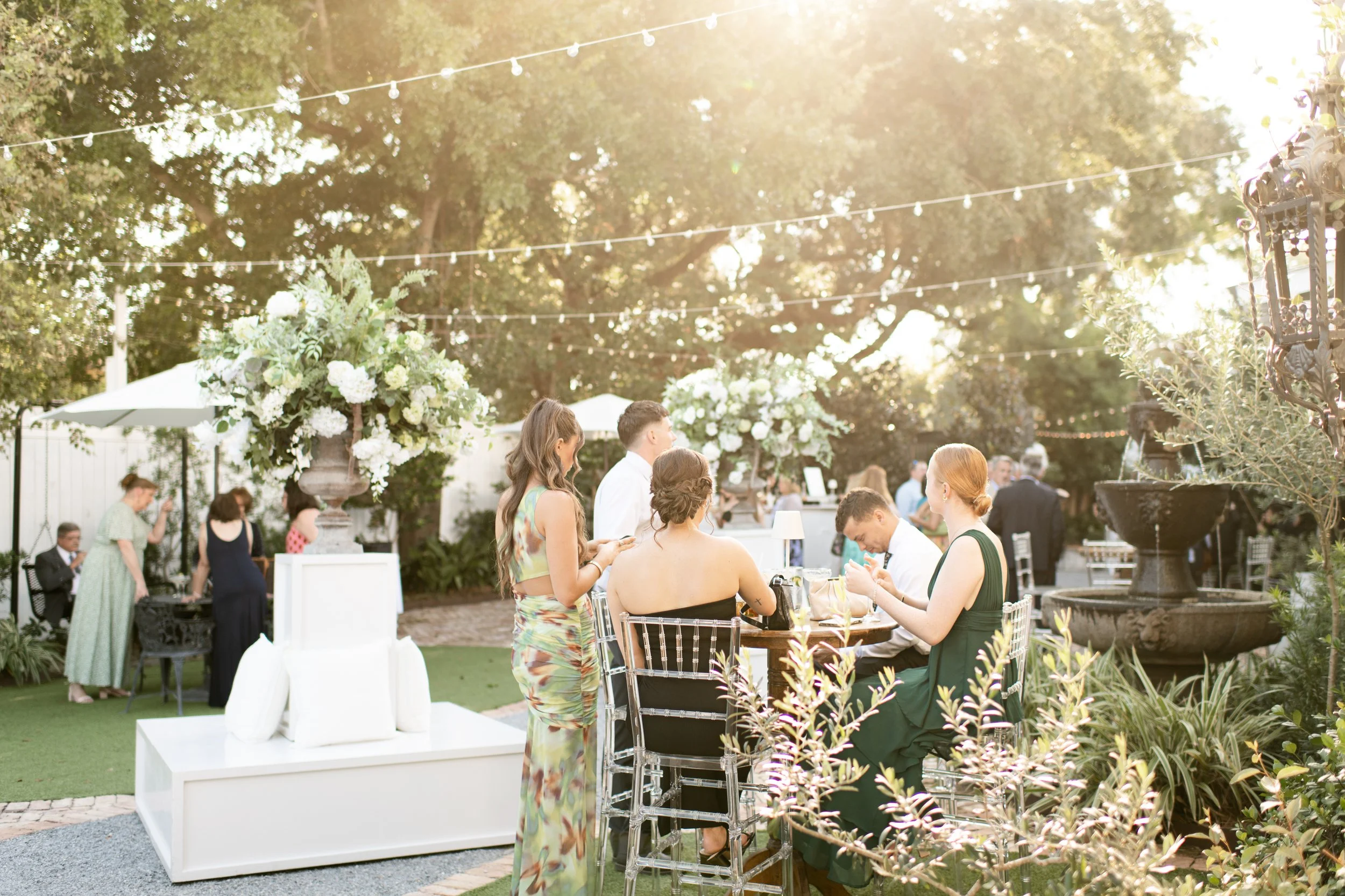 People socializing at an outdoor garden party reception, with elegant floral arrangements, string lights, and a fountain, during sunset.
