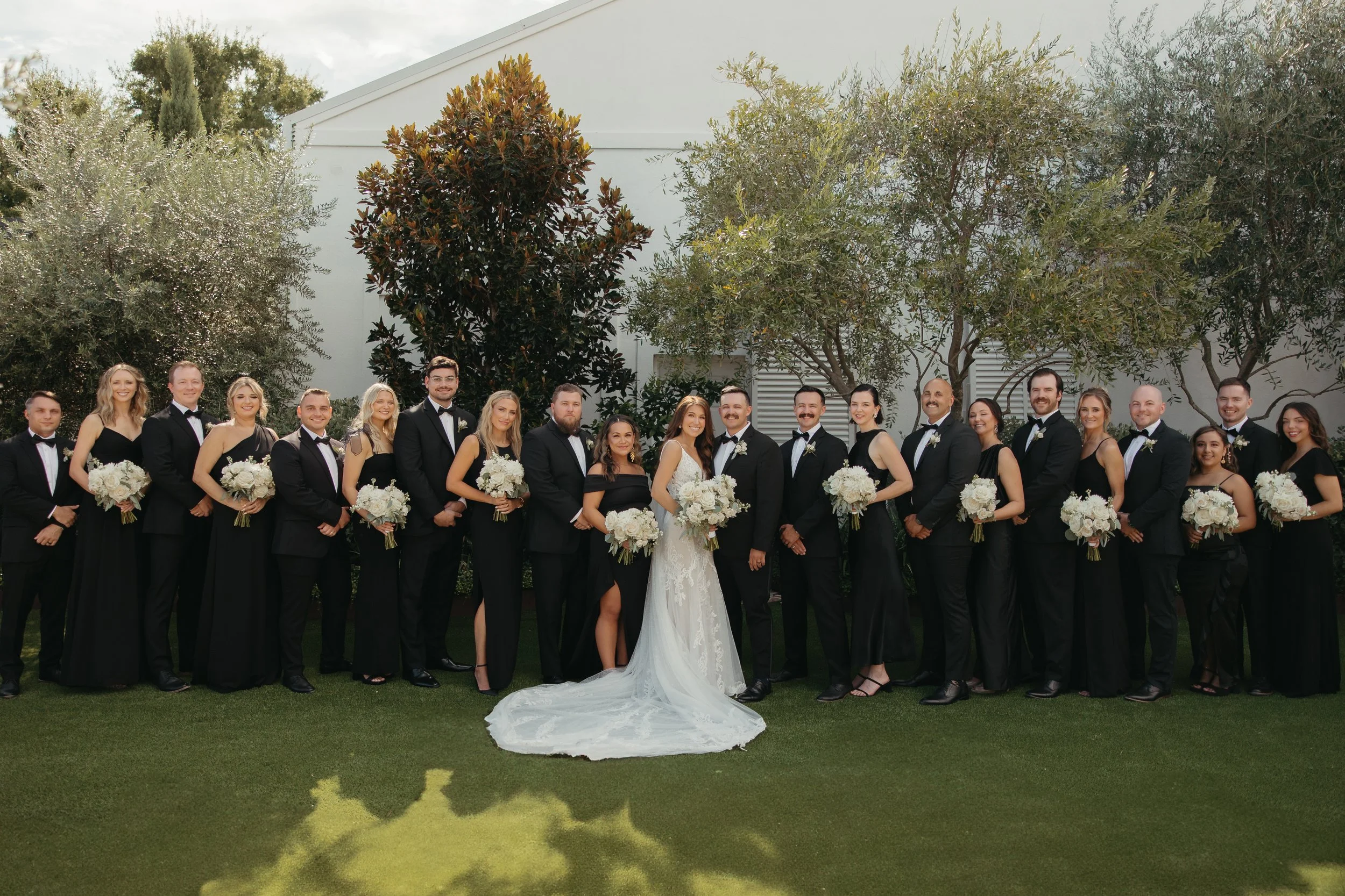 A large wedding party group photo outdoors on a grassy lawn, with bride and groom in the center, surrounded by bridesmaids and groomsmen, all dressed in formal black and white attire, holding bouquets of white flowers, with trees and a white building