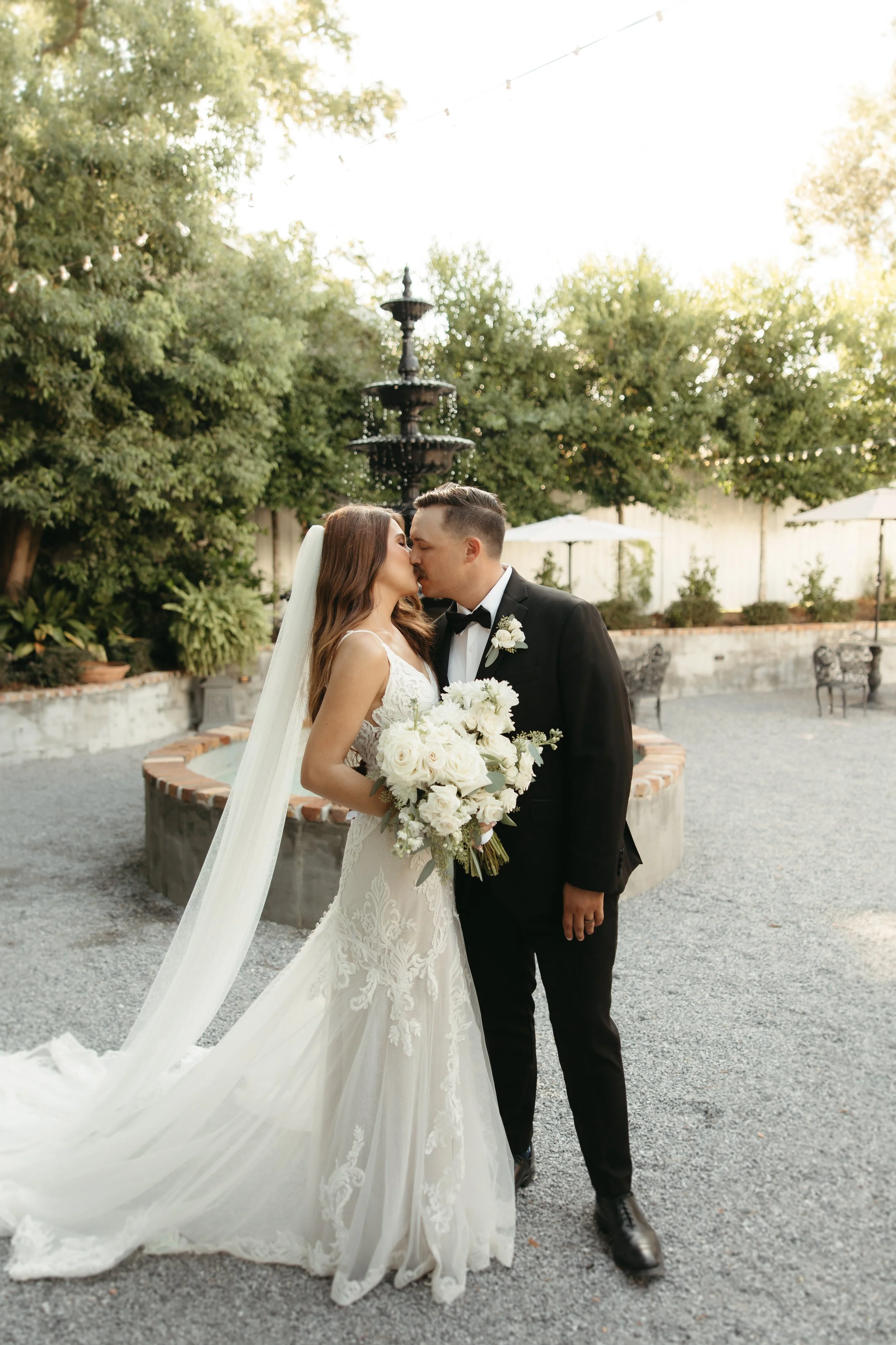 Bride and groom sharing a kiss outdoors, with the bride holding a large bouquet of white flowers, wearing a lace wedding gown and veil, and the groom in a black tuxedo with a boutonniere, in a garden setting with trees and a fountain.