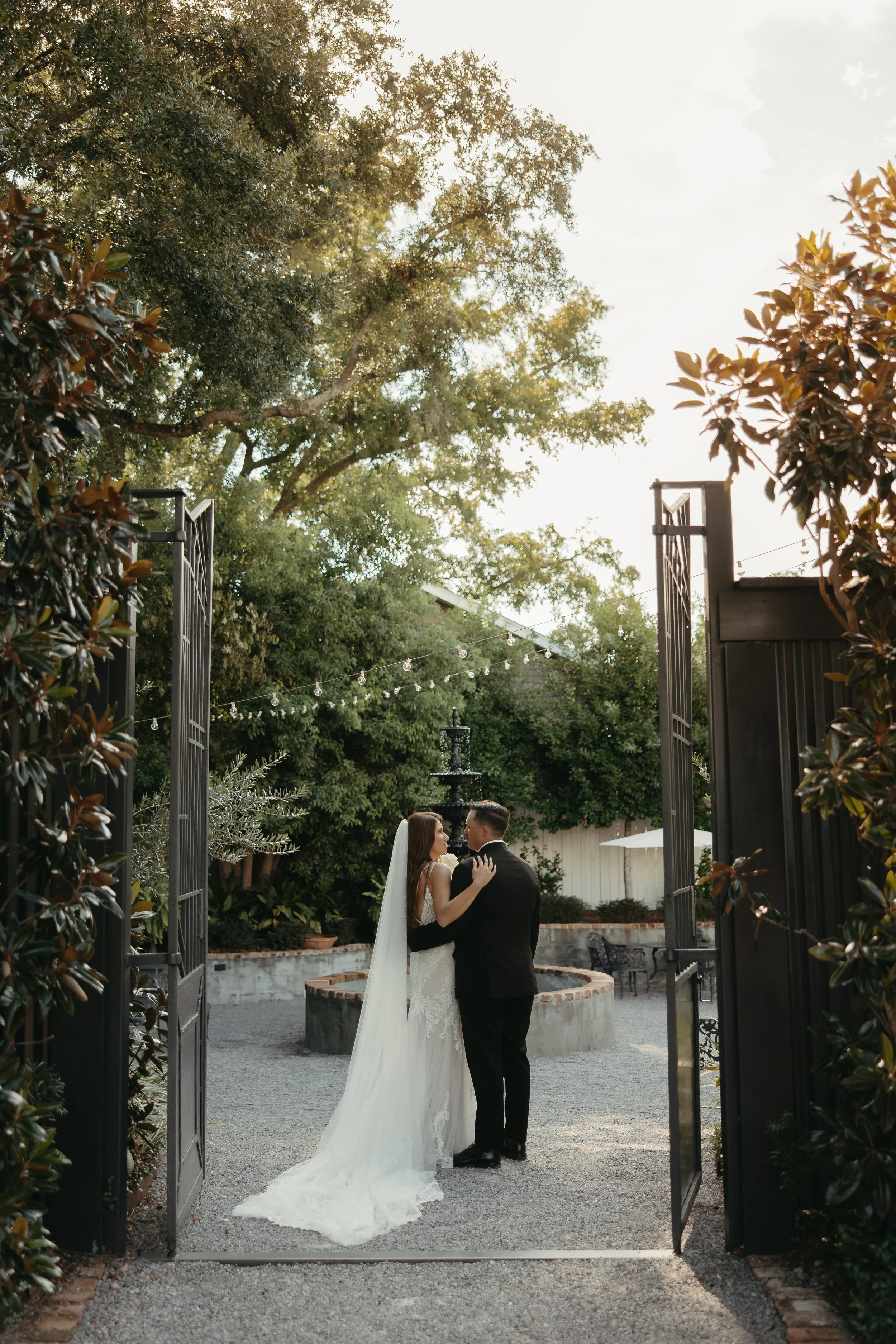A bride and groom embracing each other outside an open gate in a garden setting during sunset, surrounded by trees and string lights.
