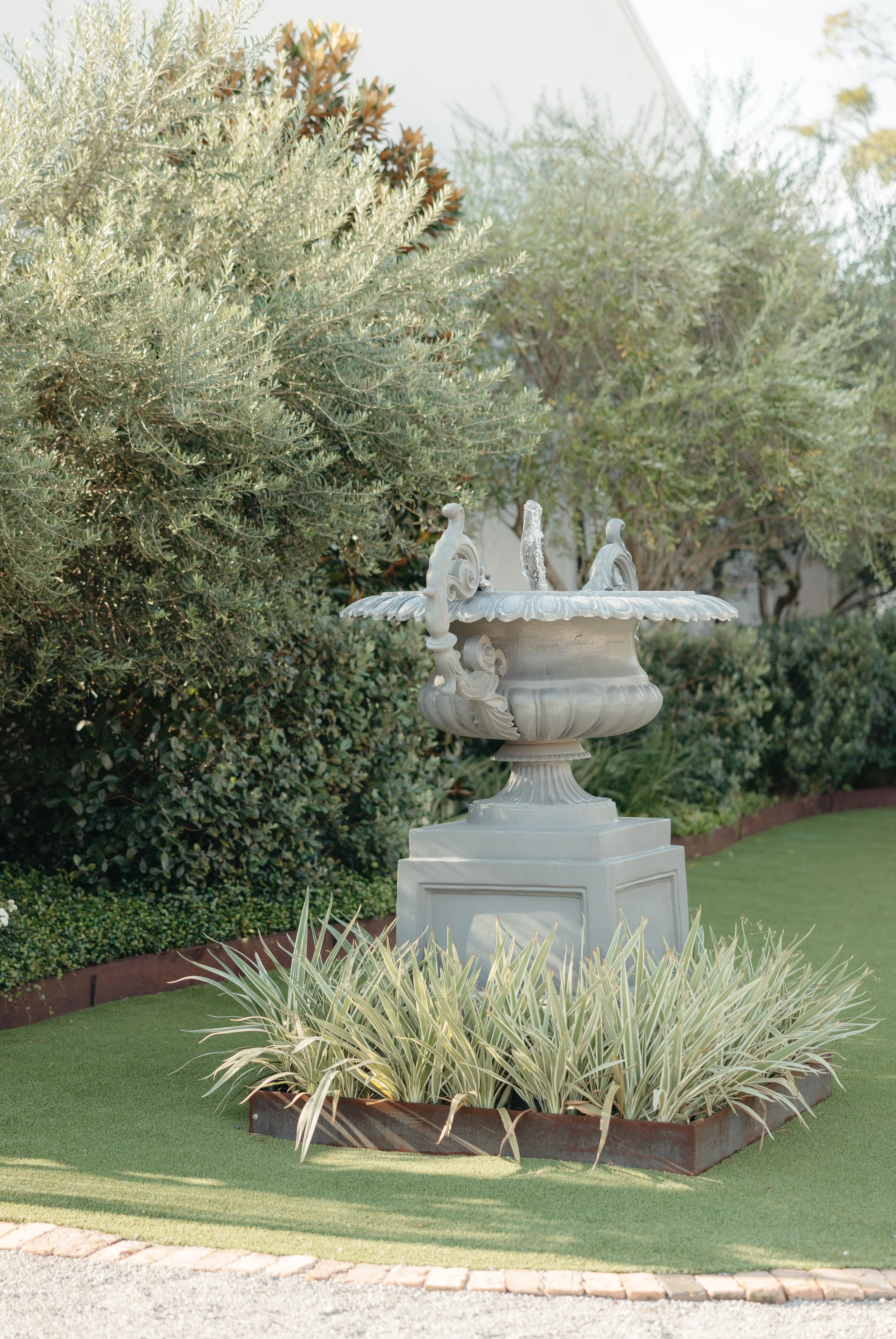 A Pensacola garden scene featuring a stone fountain with water spouting from the top, surrounded by green shrubs and plants, with a well-maintained grassy area and decorative border.