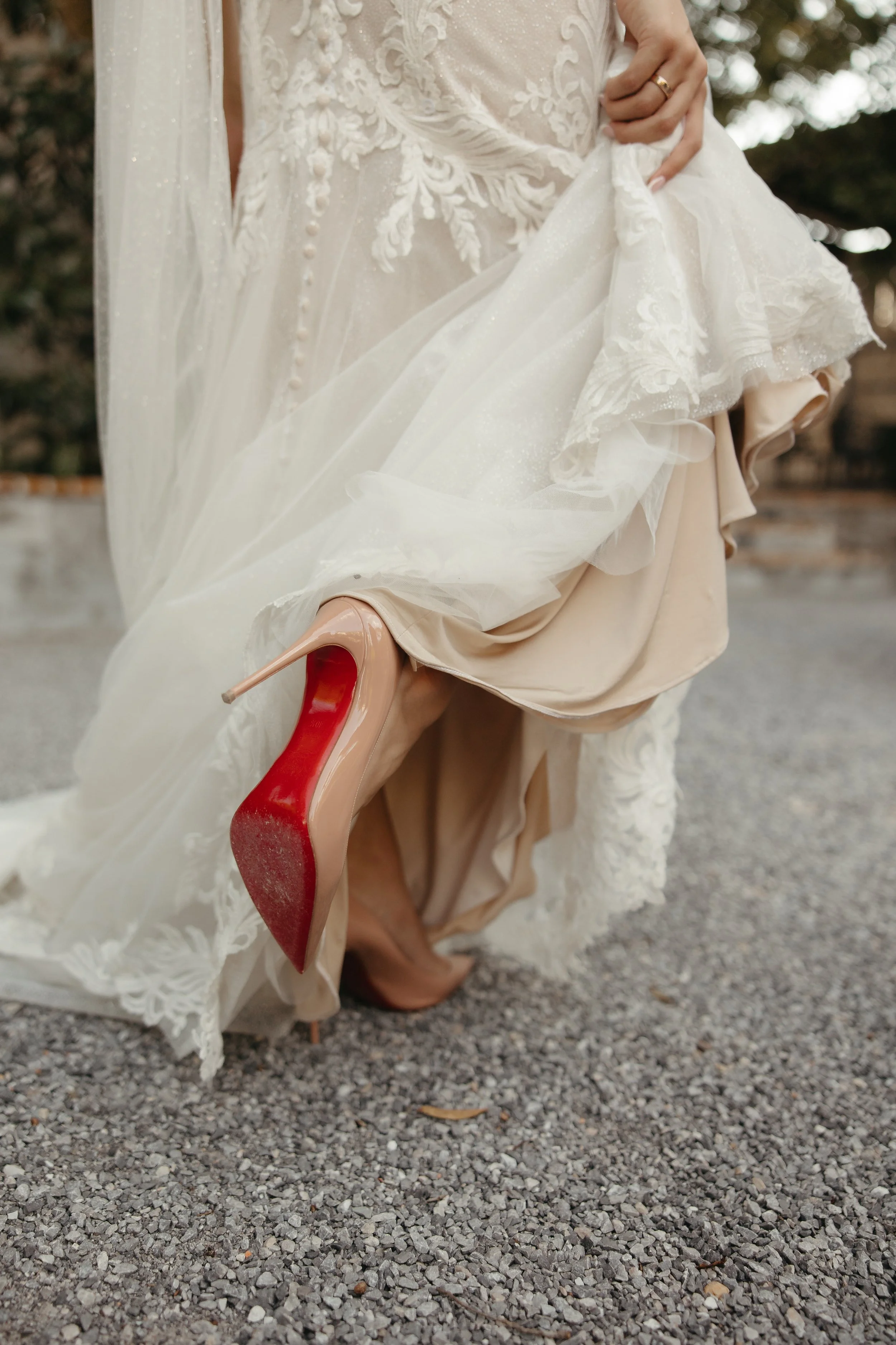 A bride lifting her wedding dress to show her nude and red-soled high heels, standing on gravel ground outdoors.