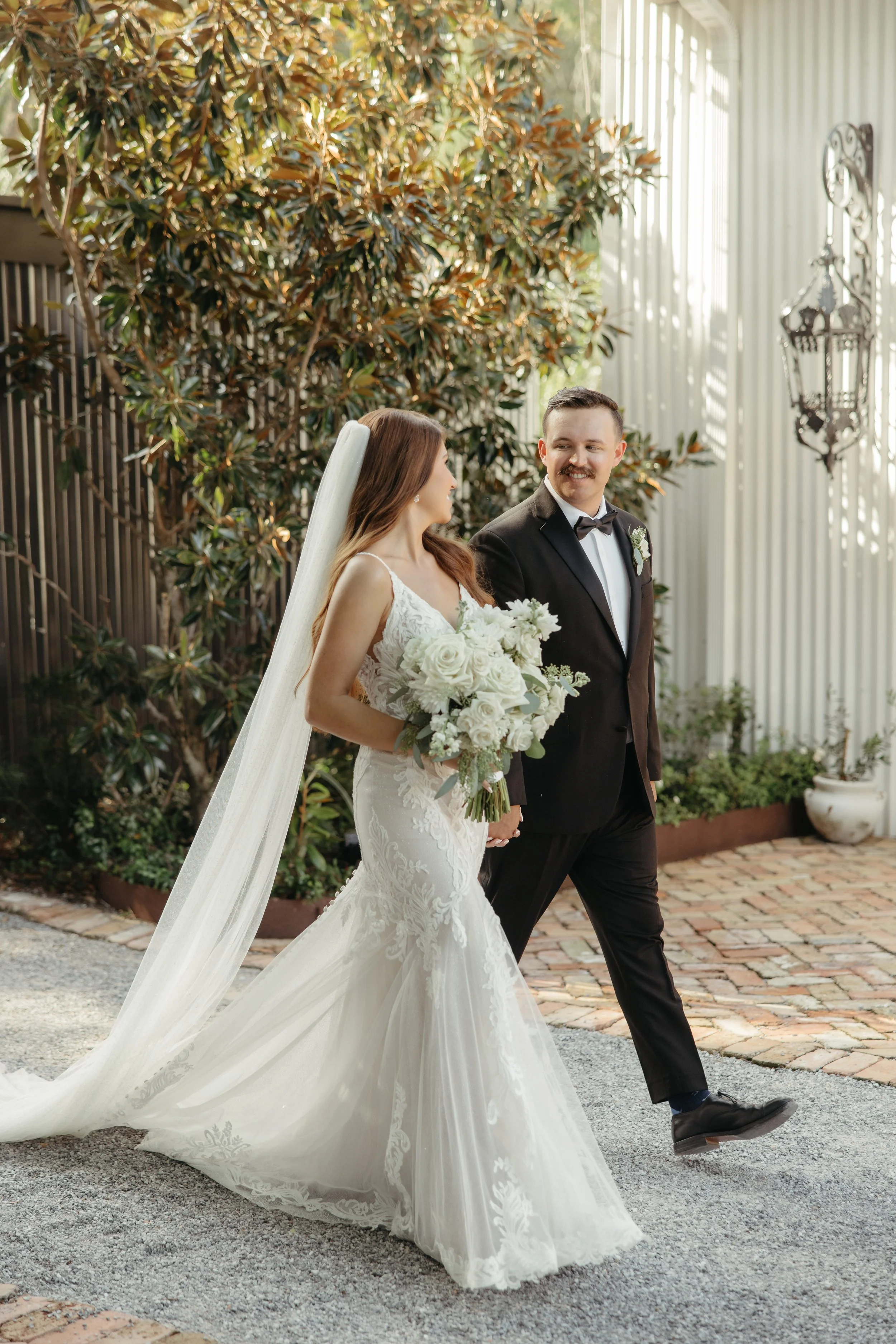 A bride and groom walking outdoors, holding hands, with the bride holding a bouquet of white flowers, wearing a white lace wedding gown with a long train and veil, and the groom in a black tuxedo with a bow tie, smiling at each other, in a garden set