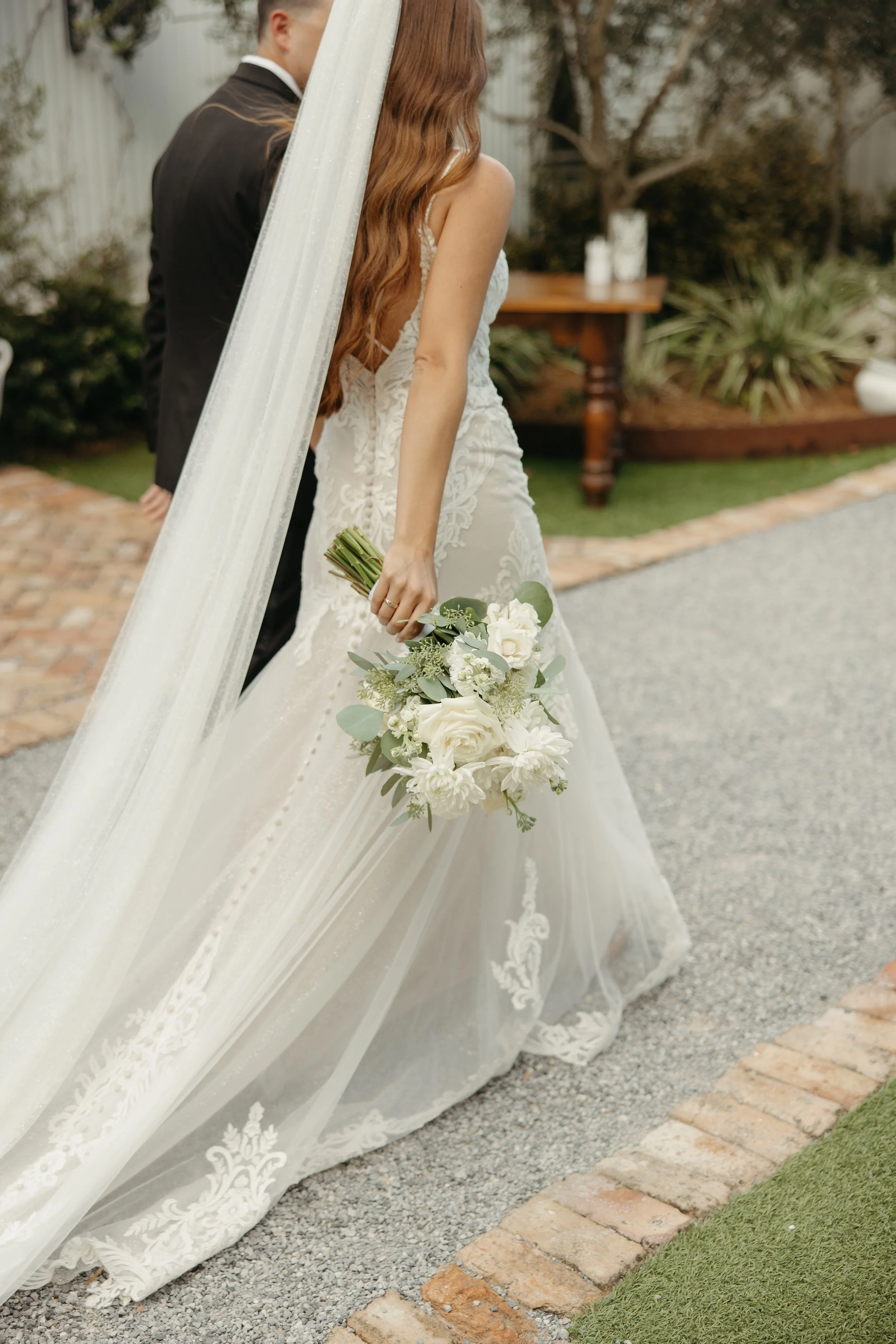 A Pensacola bride in a white lace wedding gown holding a bouquet of white flowers, standing next to a groom in a dark suit, with a veil hanging from her head, outdoors in a garden setting.