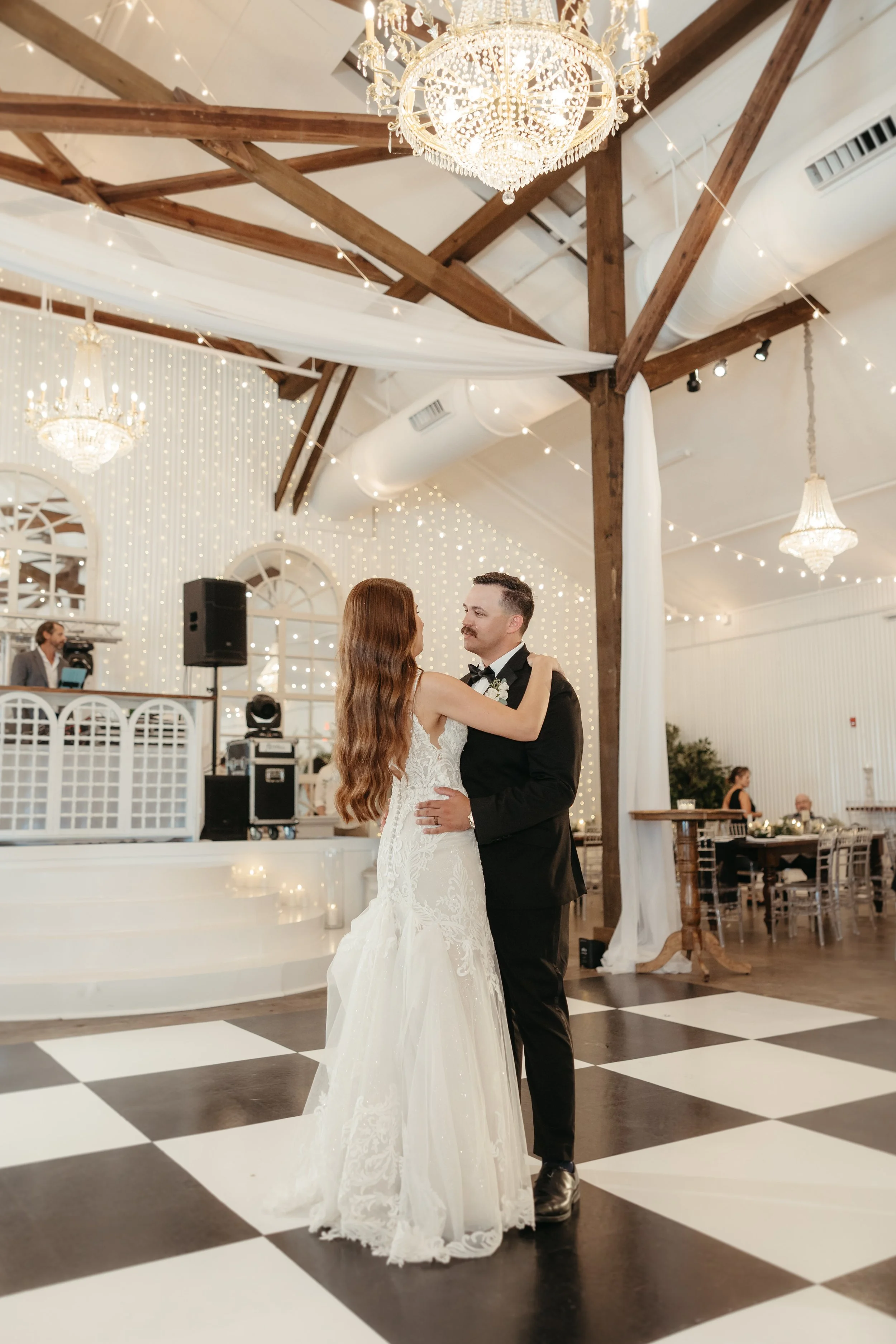 A bride and groom dancing at their wedding reception with chandeliers, string lights, and a DJ in the background.