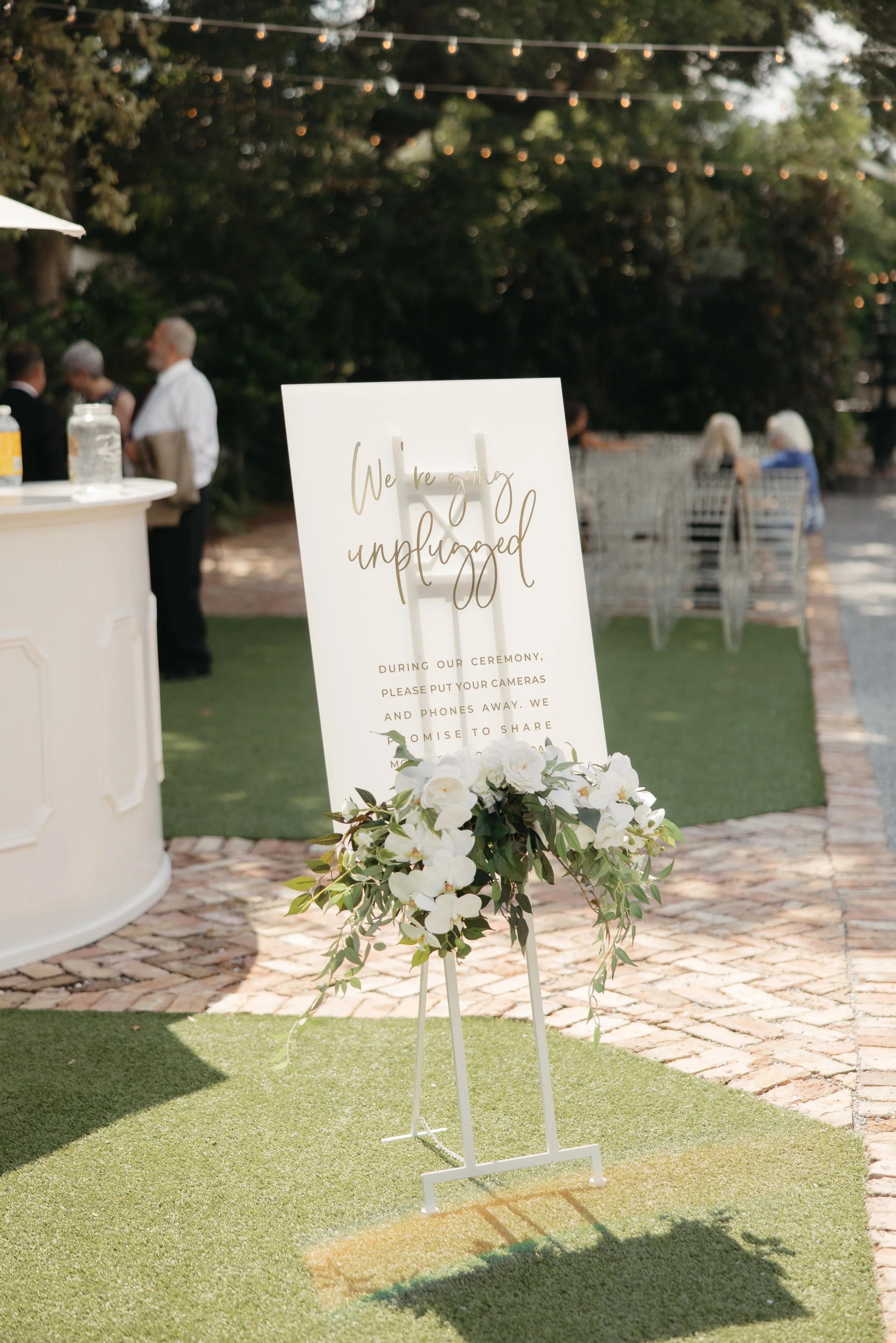 Wedding sign that reads 'We're unplugged' with floral decoration, set outdoors with guests in the background.
