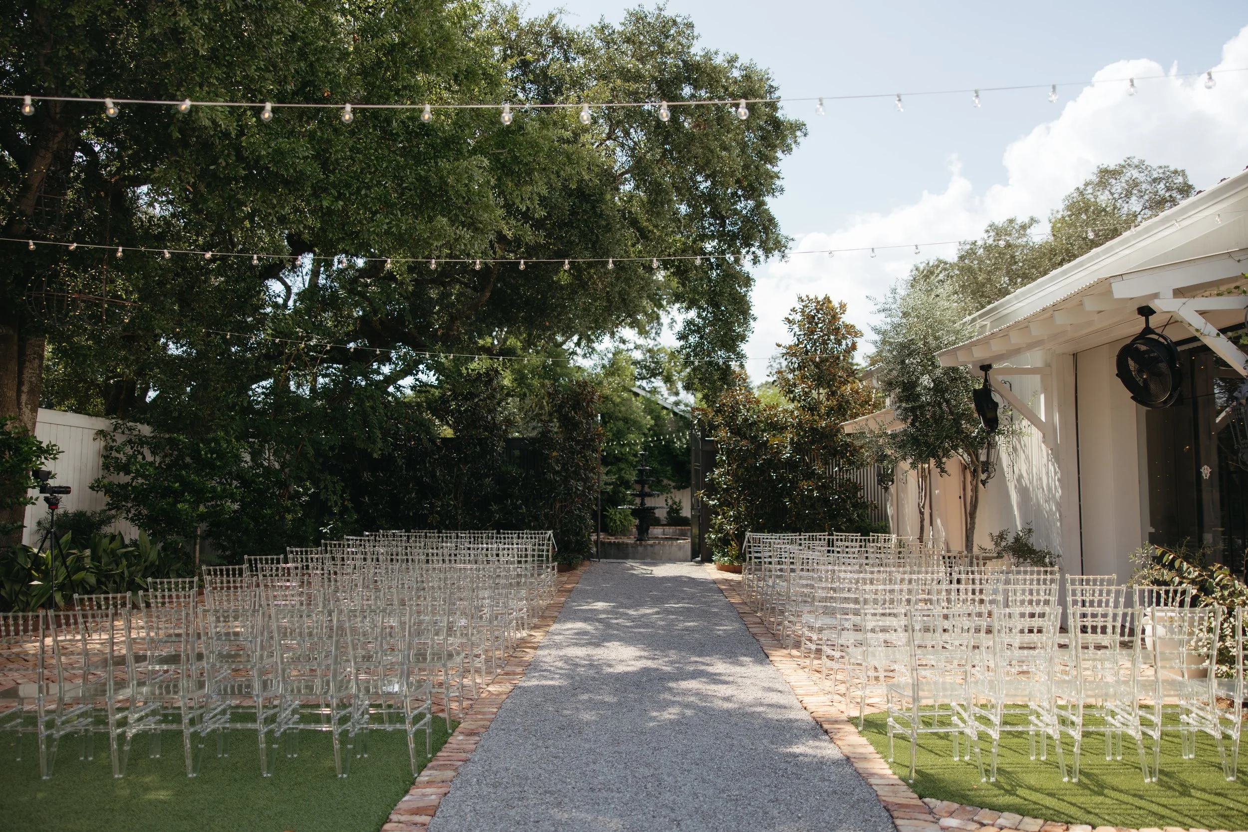 Outdoor Pensacola wedding ceremony setup with transparent chairs arranged on both sides of a gravel aisle, hanging string lights, trees, and a white building with black speakers.