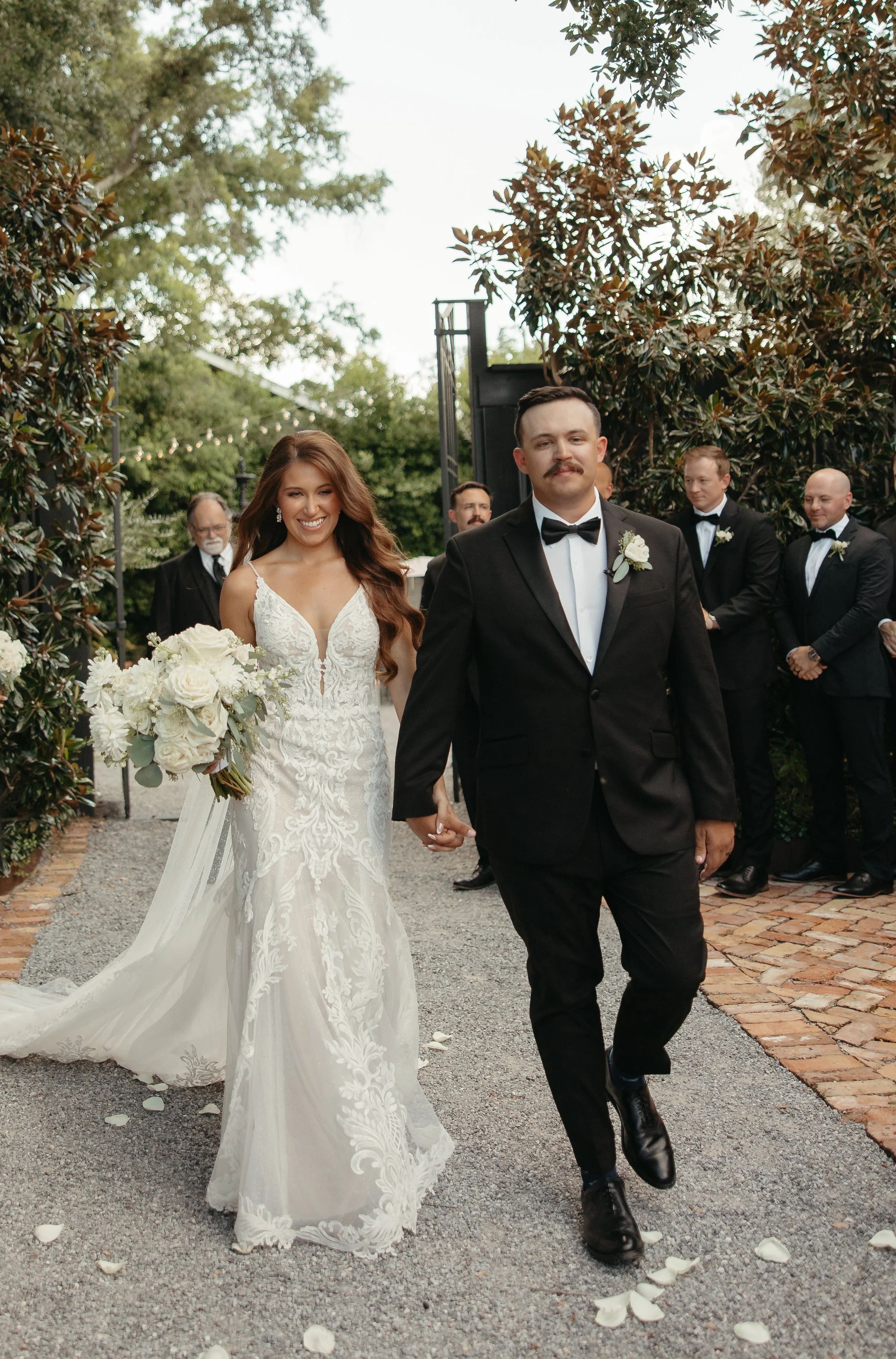 A newlywed couple walks hand in hand, smiling, during their outdoor wedding ceremony. The bride wears a white lace wedding dress and holds a bouquet of white roses and greenery. The groom wears a black tuxedo with a bow tie. Several groomsmen and an 