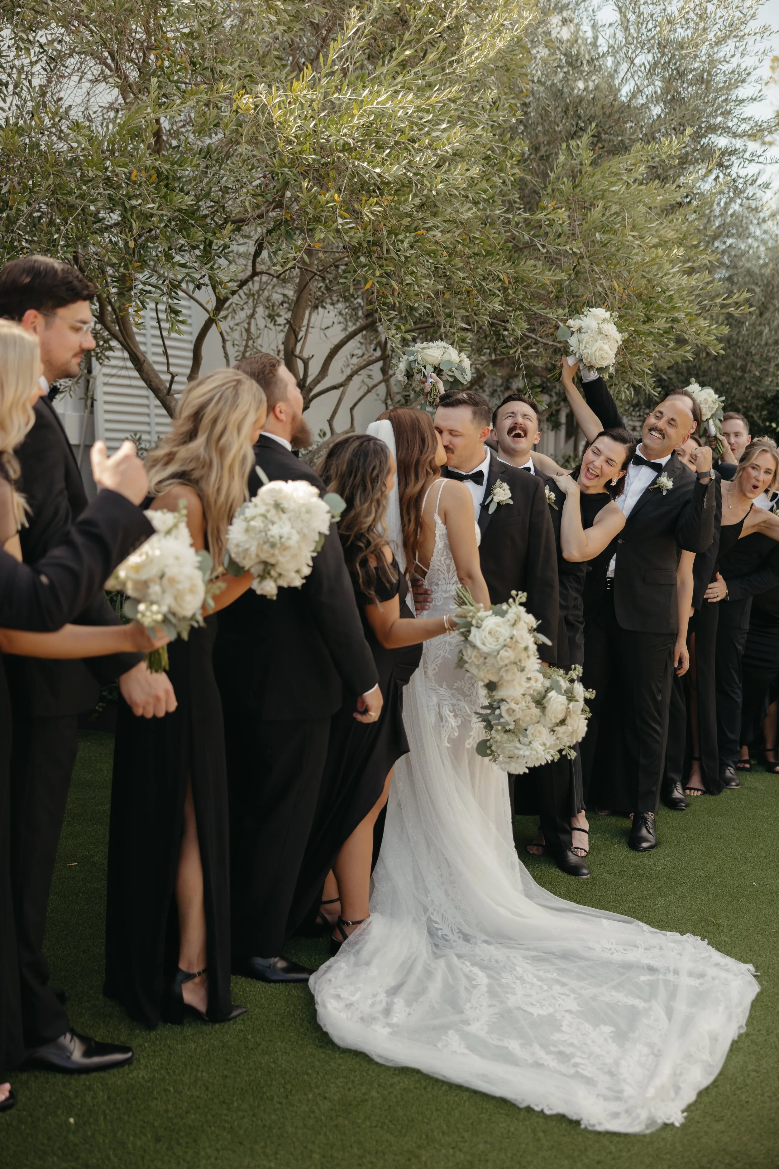 A wedding celebration with a bride and groom kissing, surrounded by friends and family, some raising their bouquets and cheering, outdoors with greenery in the background.