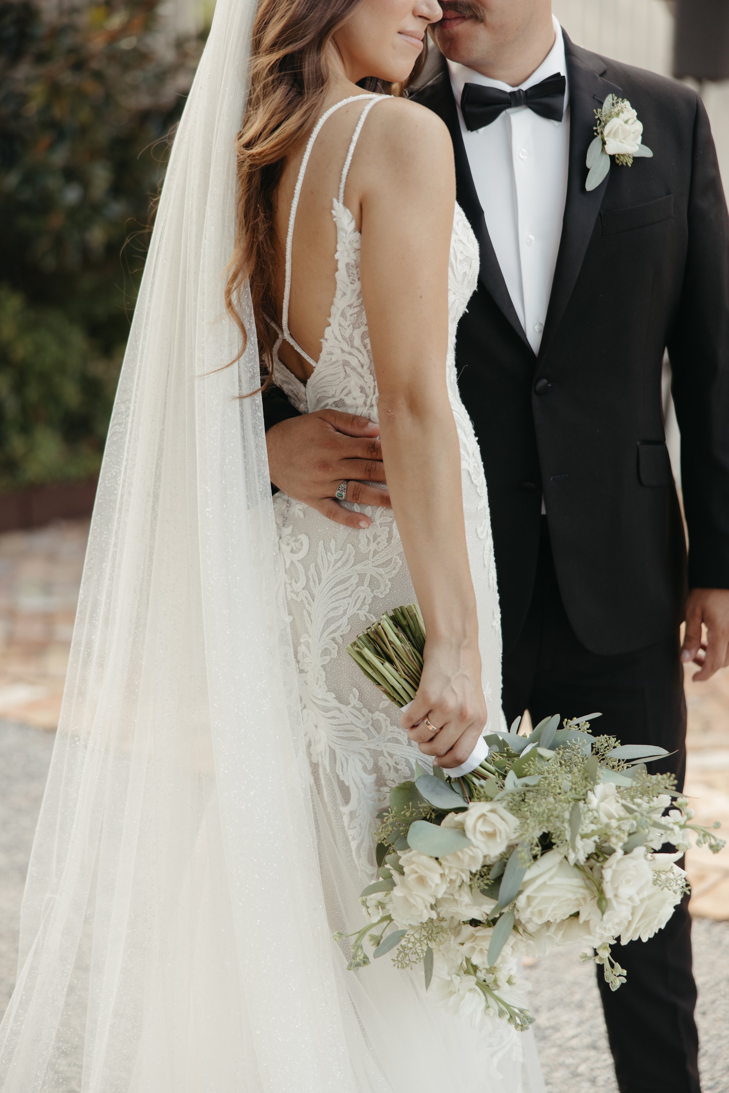 Close-up of a bride and groom on their wedding day, the bride holding a bouquet of white flowers and greenery, the groom wearing a black tuxedo with a white boutonniere, embracing outdoors.