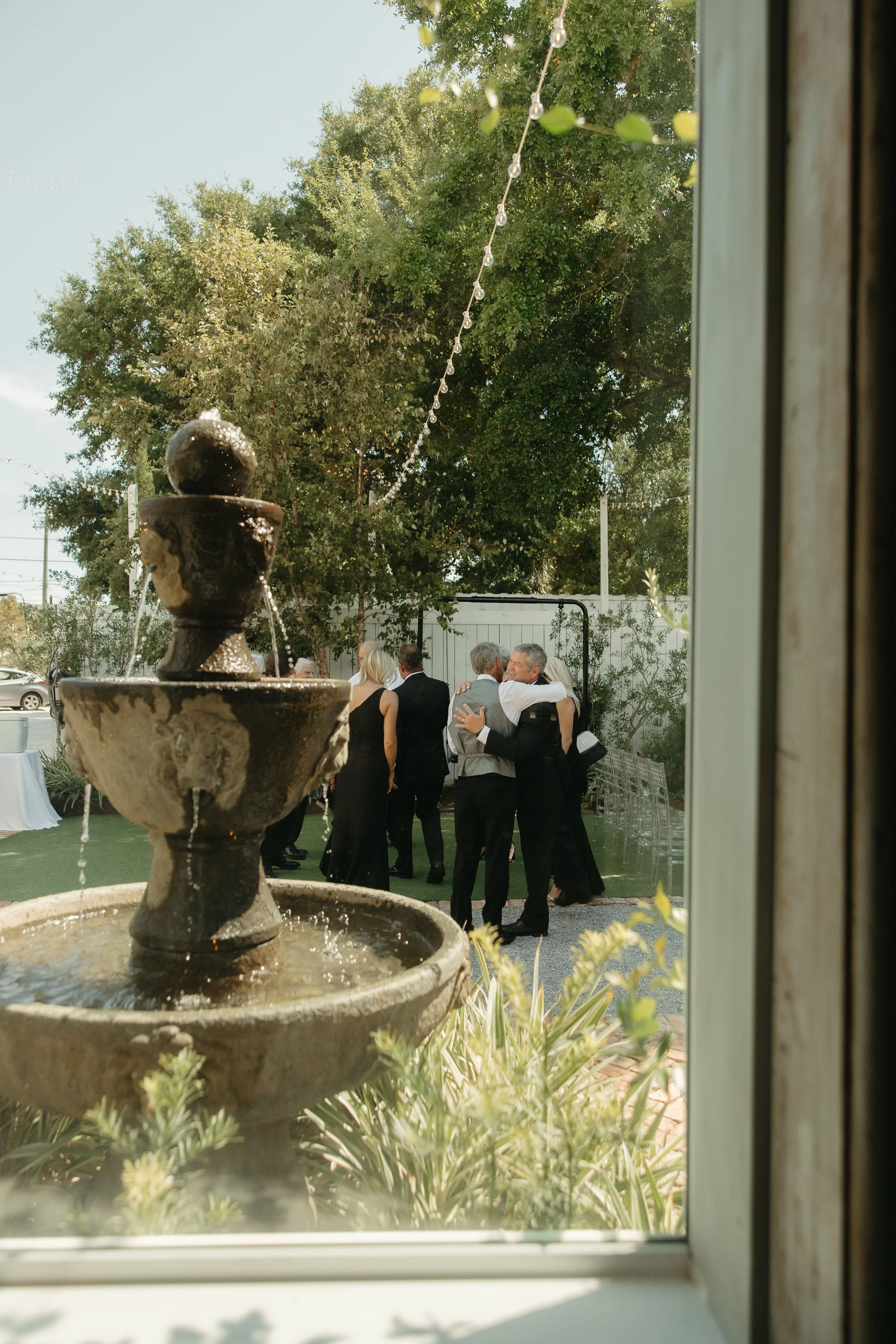 People dancing and socializing outdoors at a gathering or celebration, as seen through a window with a fountain in the foreground.
