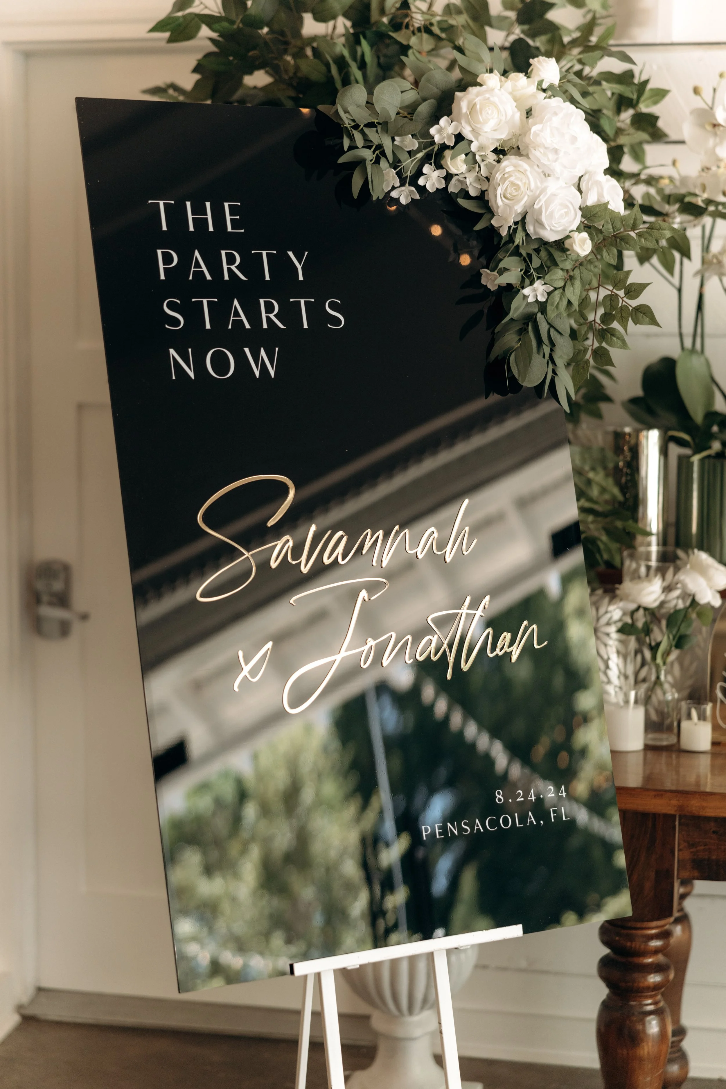 A black wedding sign on an easel with white and gold cursive and block text reading 'The party starts now' and 'Savannah x Jonathan', decorated with white flowers and greenery, with a background of a decorated venue in Pensacola, Florida.