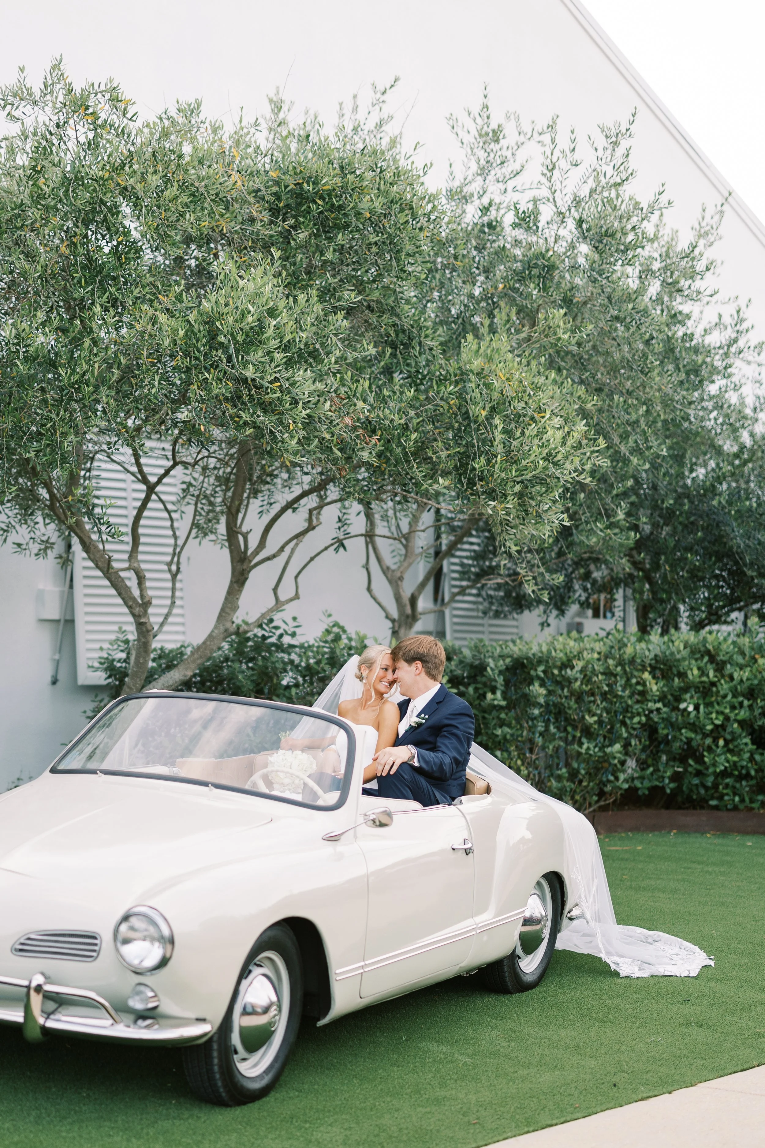 A bride and groom sitting in a vintage white convertible car, smiling and close together, on a green lawn with trees and a white building in the background.