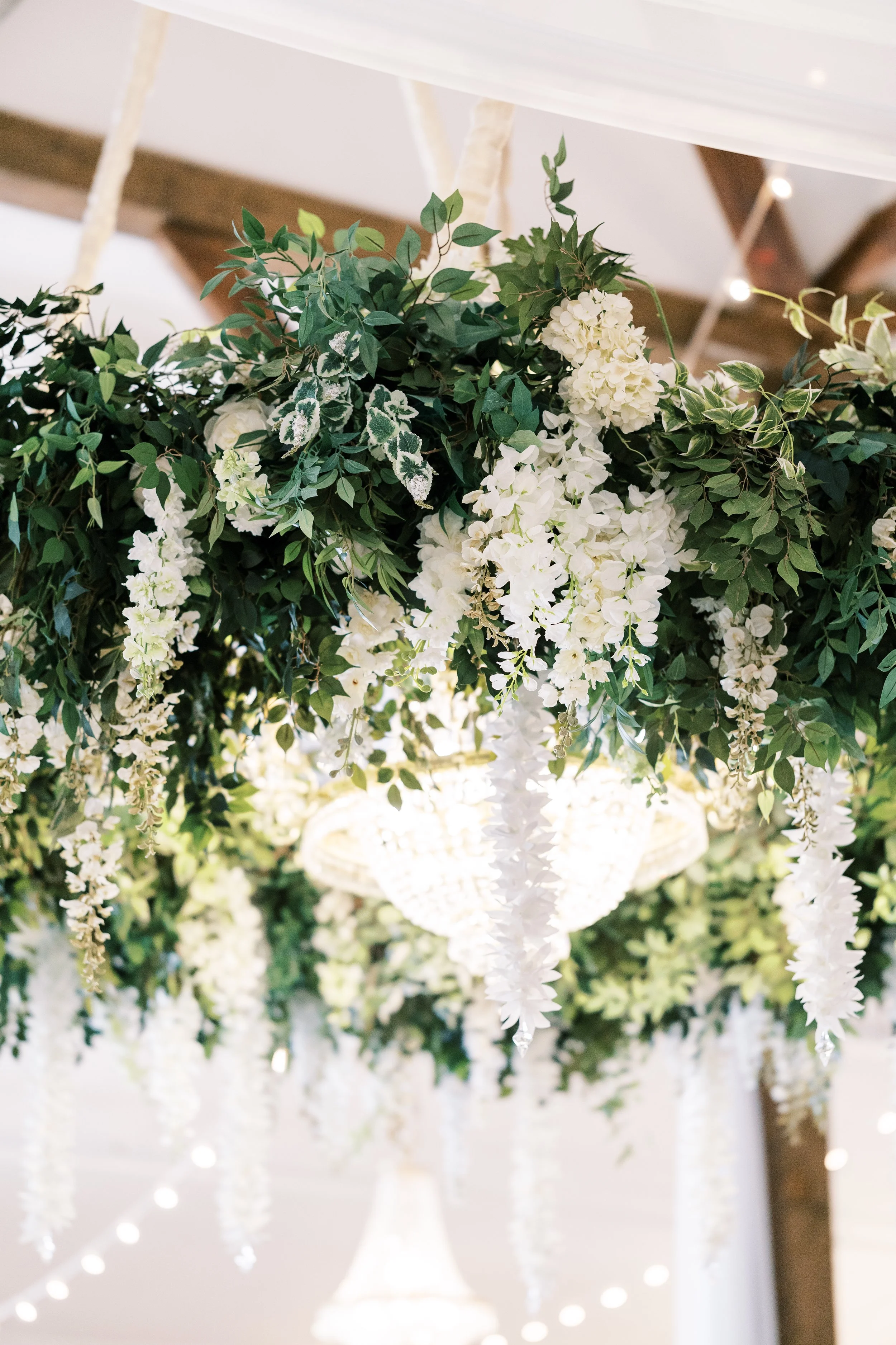 Hanging floral centerpiece with white flowers and green leaves over a chandelier in a decorated wedding venue.