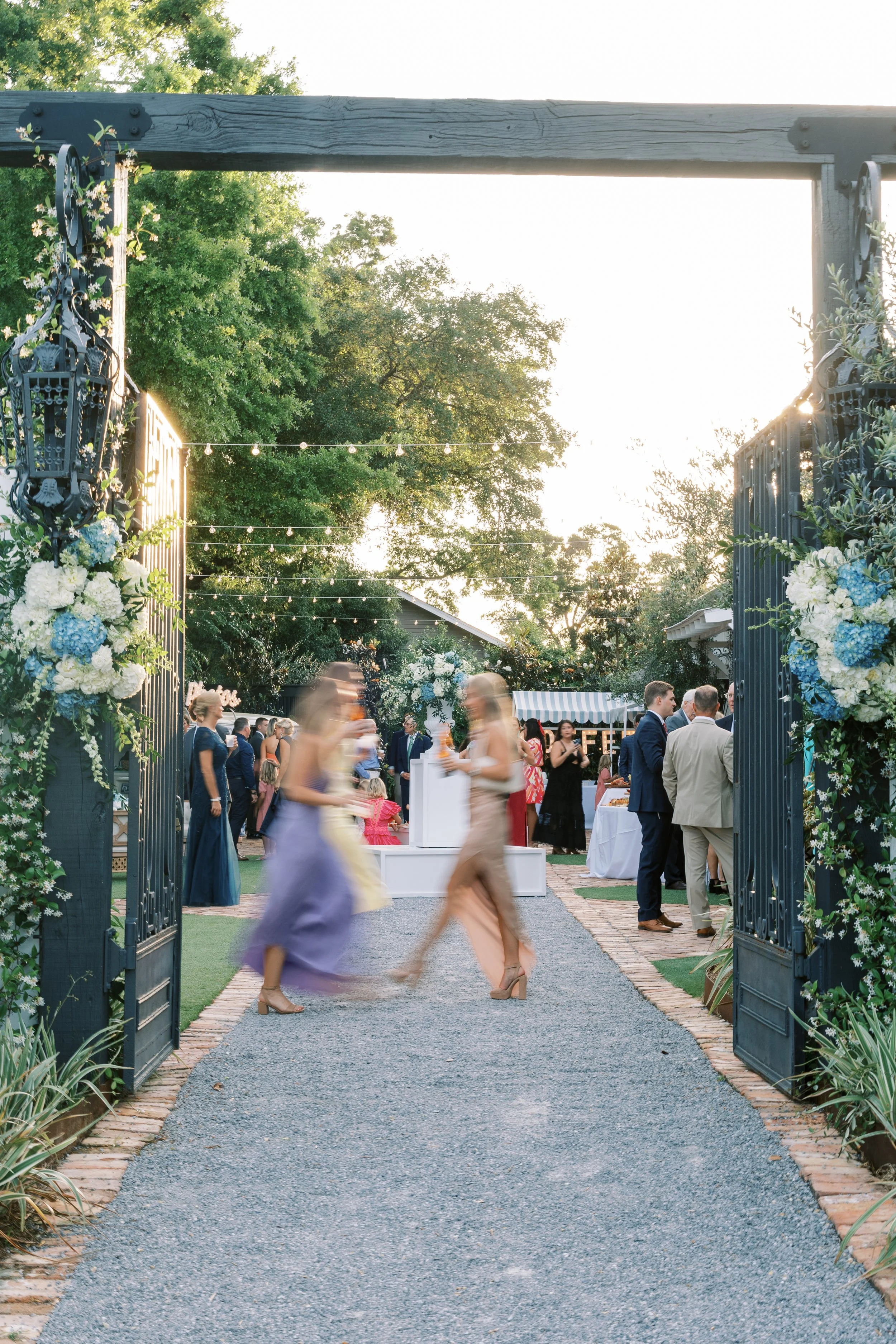 Pensacola Wedding reception scene viewed through an open gate with floral decorations. Guests are mingling outdoors under string lights during sunset.