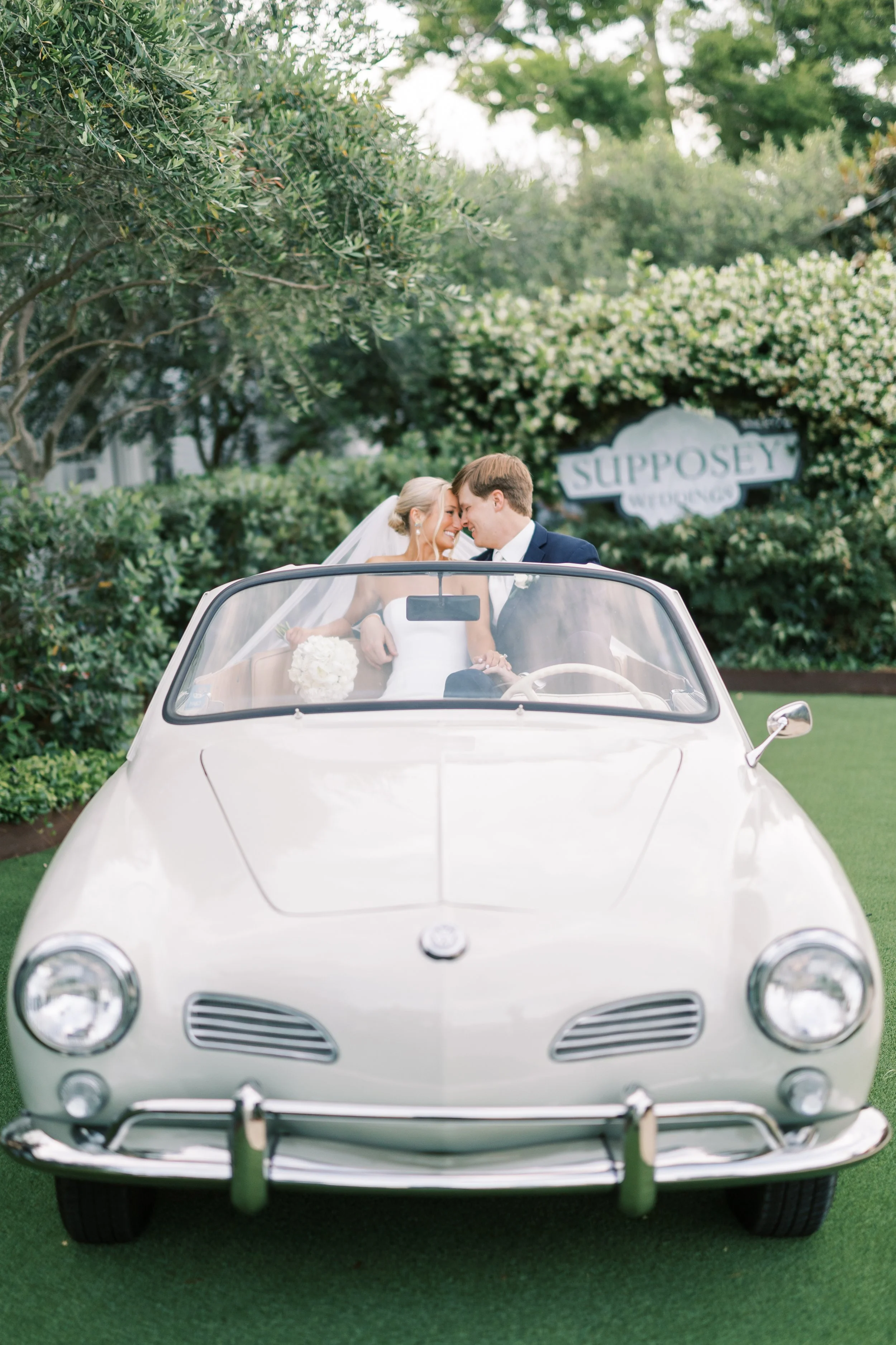 A bride and groom sit in a vintage white convertible car, smiling and leaning close to each other on their wedding day, with lush green trees and a sign that reads 'Supposey' in the background.