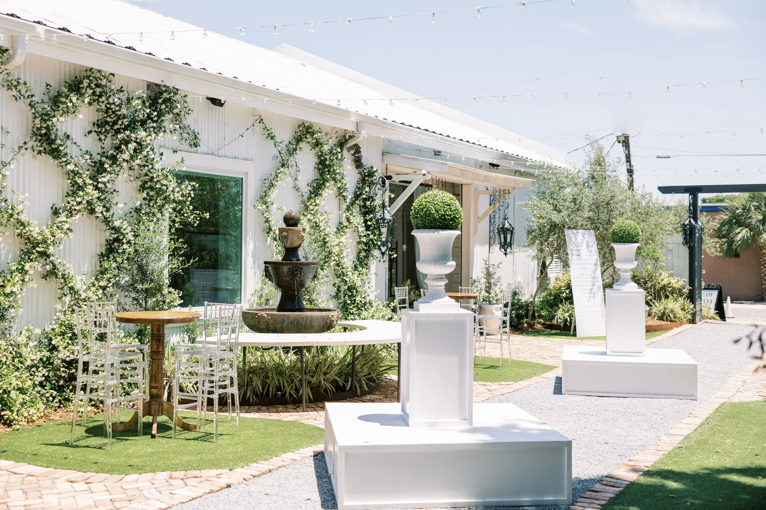 Outdoor garden setup with white pedestals topped with green potted plants, a fountain, tables, white chairs, and decorative lighting under a clear sky.