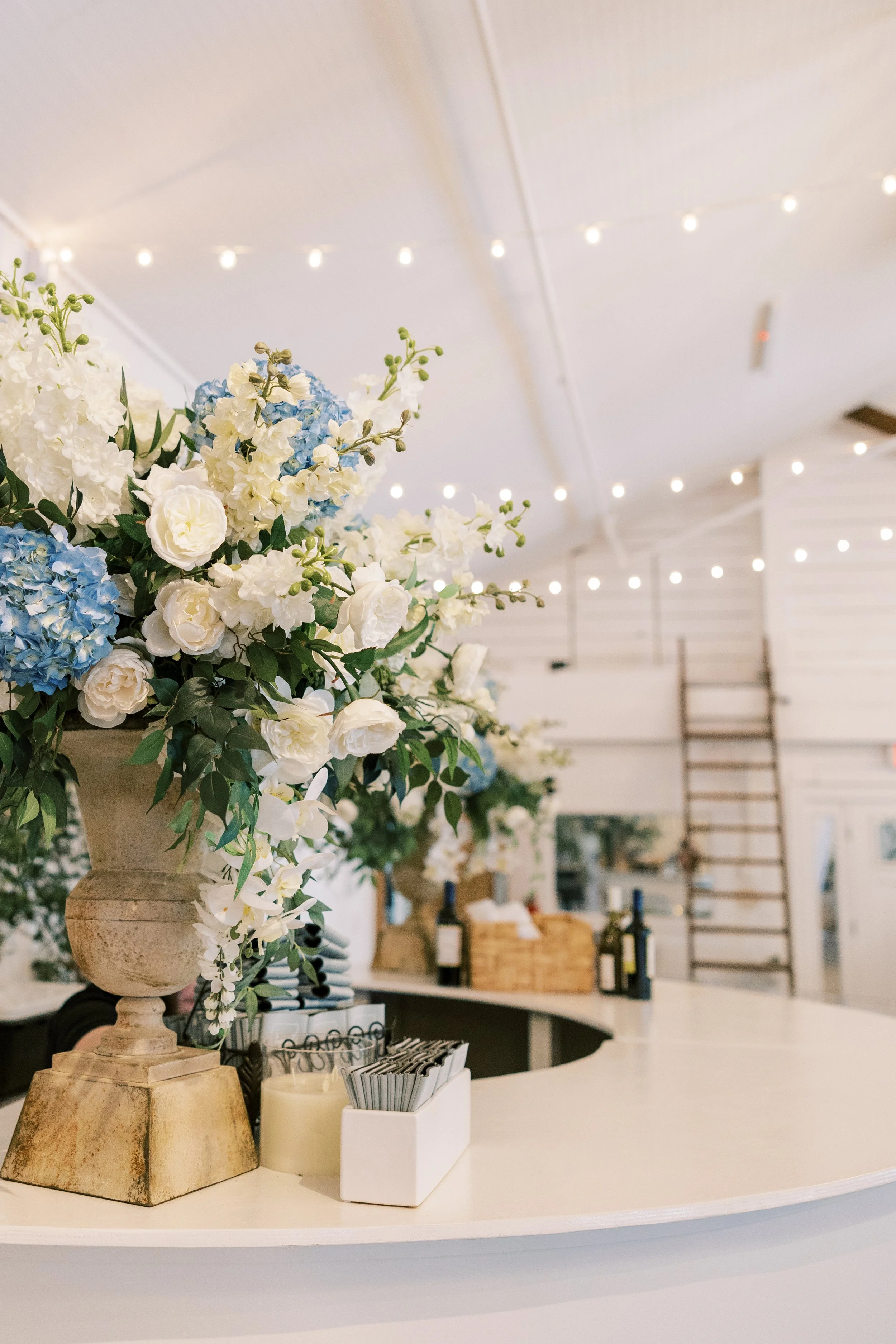 Elegant reception or bar area decorated with a large floral arrangement of white and blue flowers in a stone vase, with string lights hanging from the ceiling and wine bottles and other items on the counter behind.
